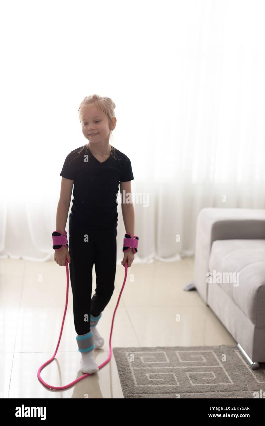 Happy little girl jumping over the rope at home during the isolation. The bright sun illuminates the room. Stock Photo