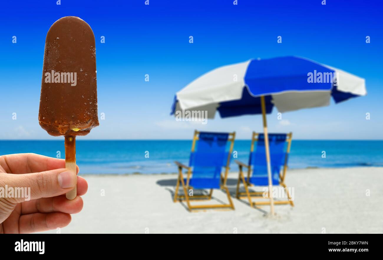 hand holding a fresh chocolate flavor popsicle in front of blurred sun loungers and a beach umbrella on silver sand Stock Photo