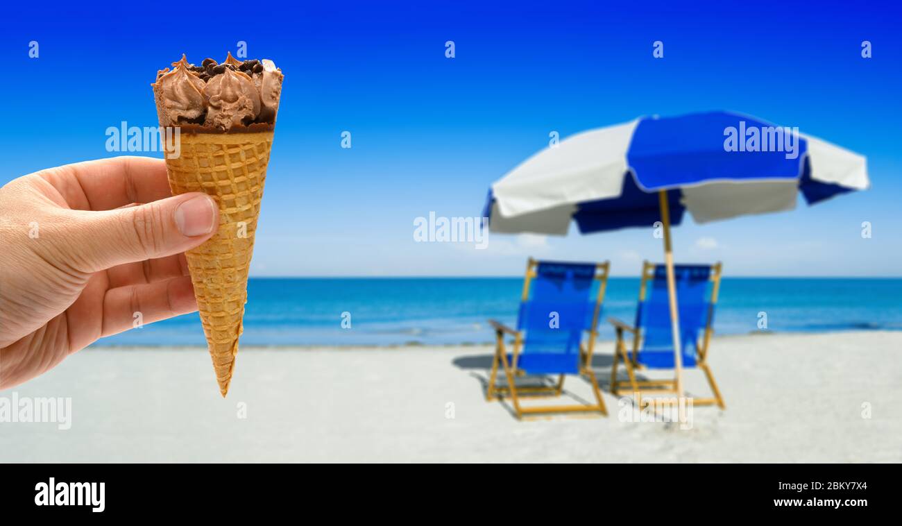 hand holding a fresh chocolate flavor ice cream cone in front of blurred sun loungers and a beach umbrella on silver sand Stock Photo