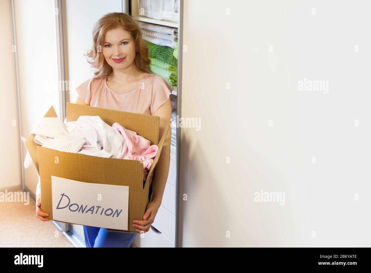 A girl stands near the wardrobe and holds a box with things. Clothes for donation. Stock Photo