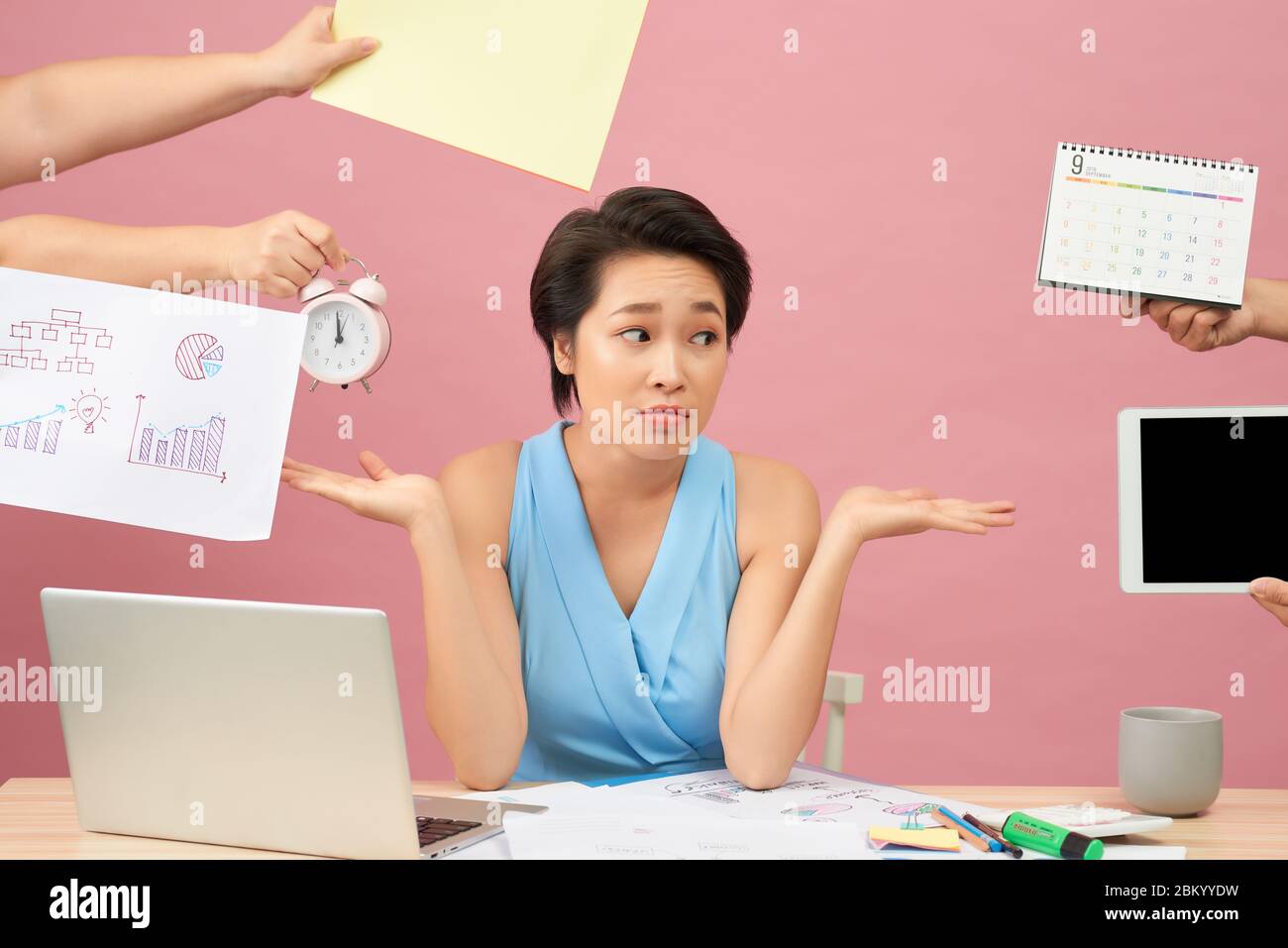 Overworked young employee refuses all things, frowns face in annoyance, sits at desktop with paper documents and notepad, isolated over pink backgroun Stock Photo