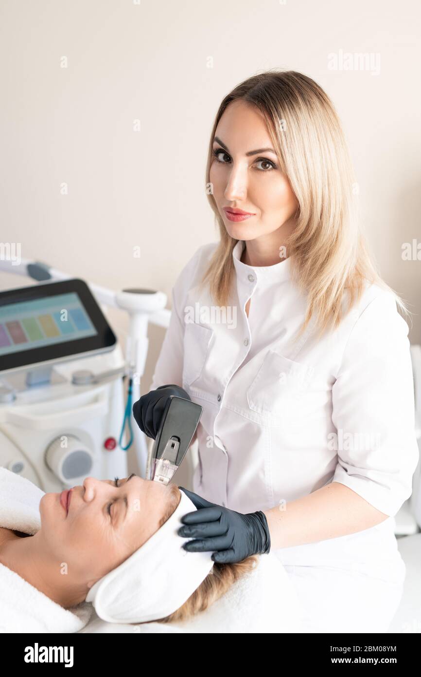 Portrait of content blond-haired beautician doing skin treatment procedure to mature woman with facial machine Stock Photo