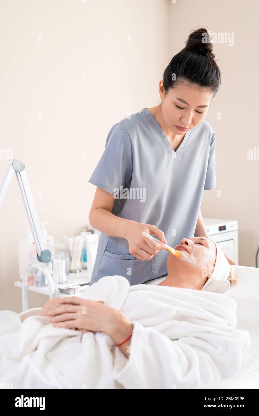 Asian beautician in scrubs applying facial mask on mature clients face using brush in beauty