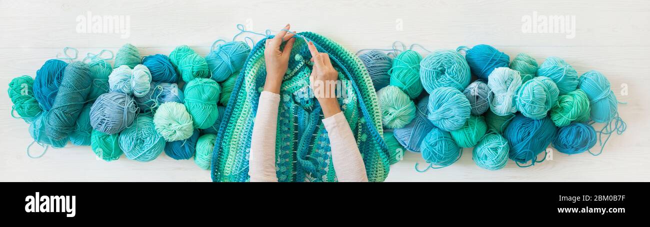 Women's hands are large. Woman crochets. Yarn of green, turquoise, aquamarine and blue colors. White wood long background. Stock Photo