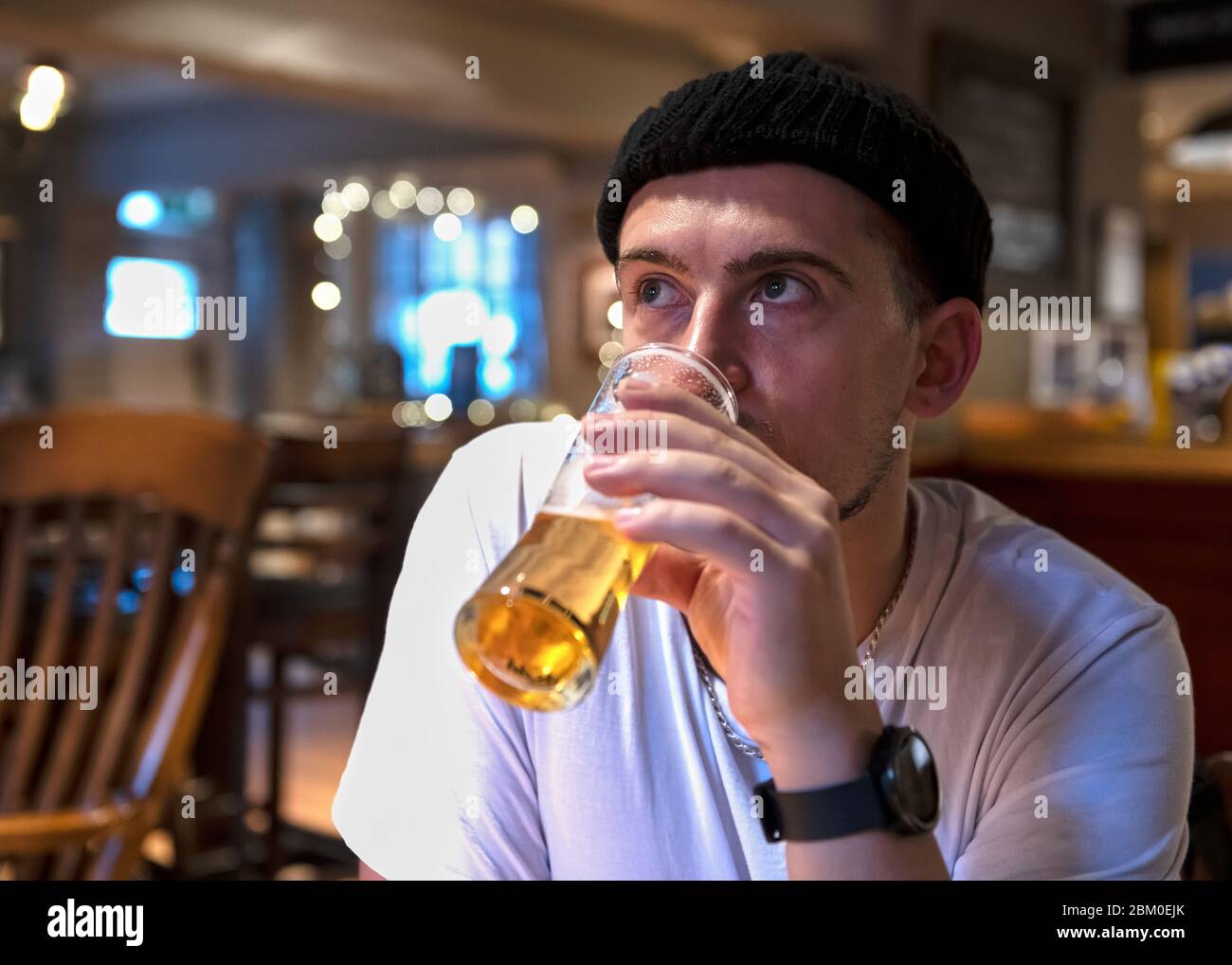 Pensive millennial male in white shirt and warm black hat watching TV while resting at table alone and drinking fresh beer from glass against blurred Stock Photo