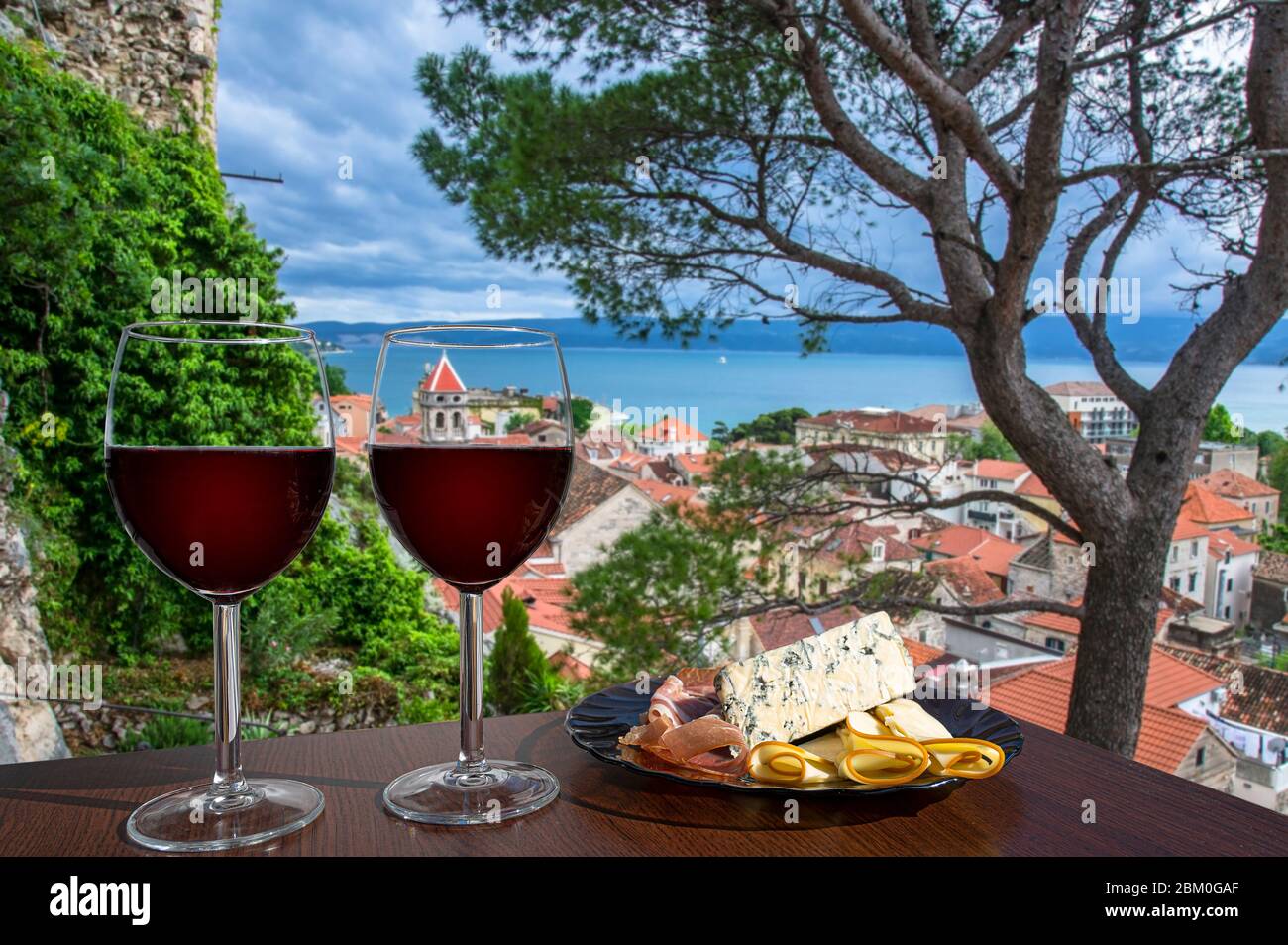 Two glasses of red wine with charcuterie assortment with view of Croatian town Omis with red roofs and blue sea. Glass of red wine with snacks. Stock Photo