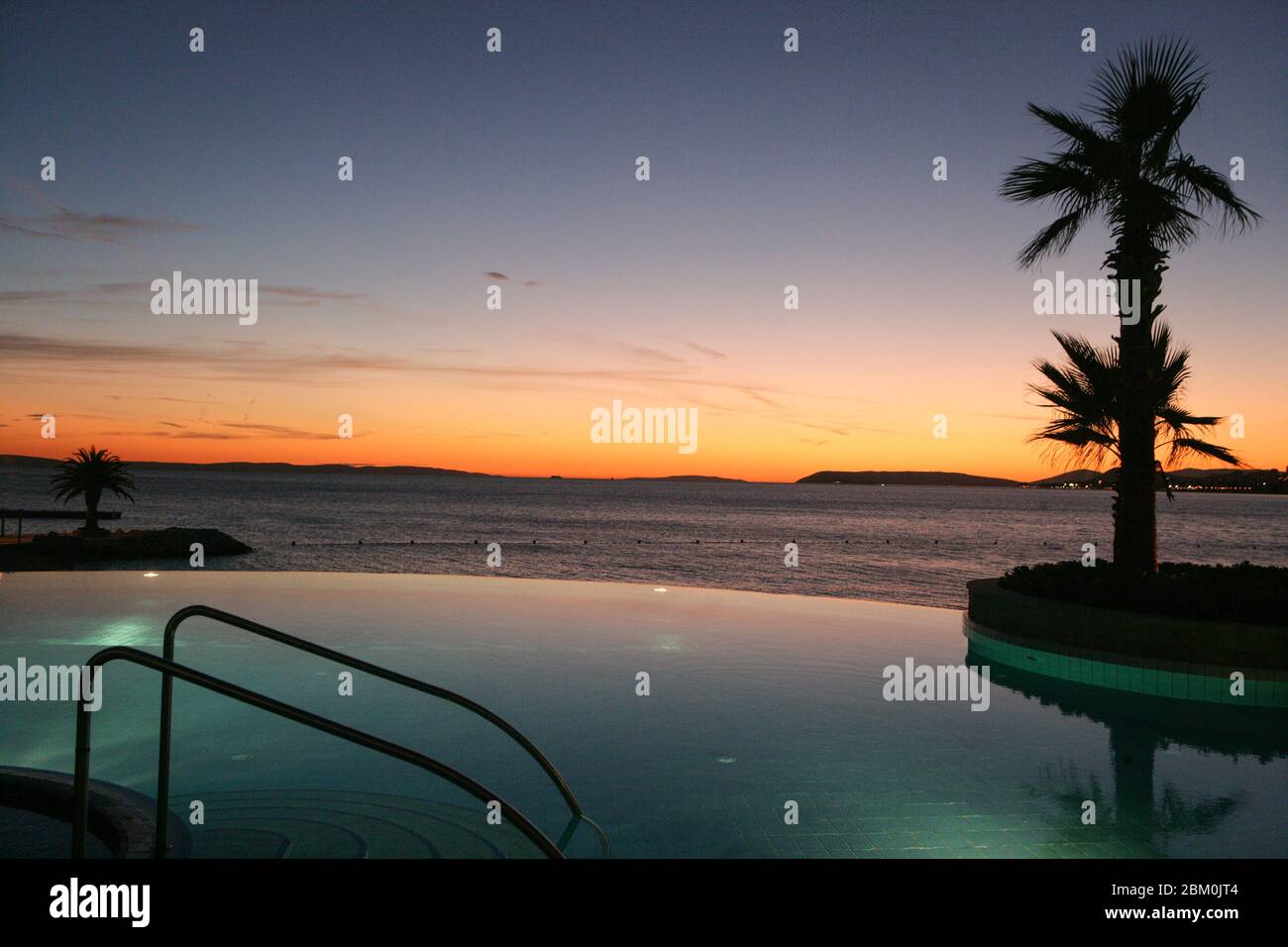 A swimming pool and a palm with the sea and the sunset in the background Stock Photo