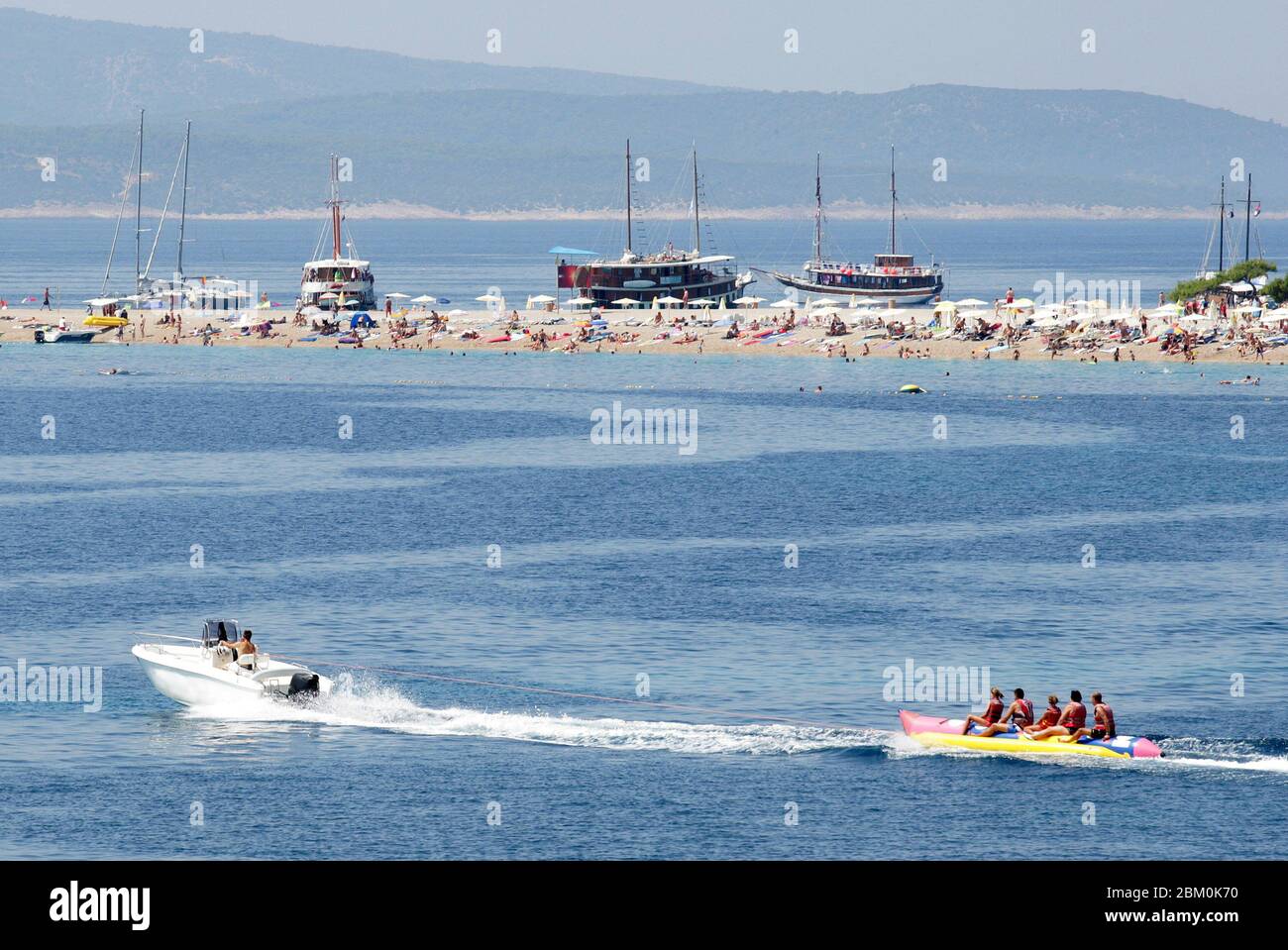 A speedboat towing people on a banana shaped float in front of a sandy beach in Bol Stock Photo