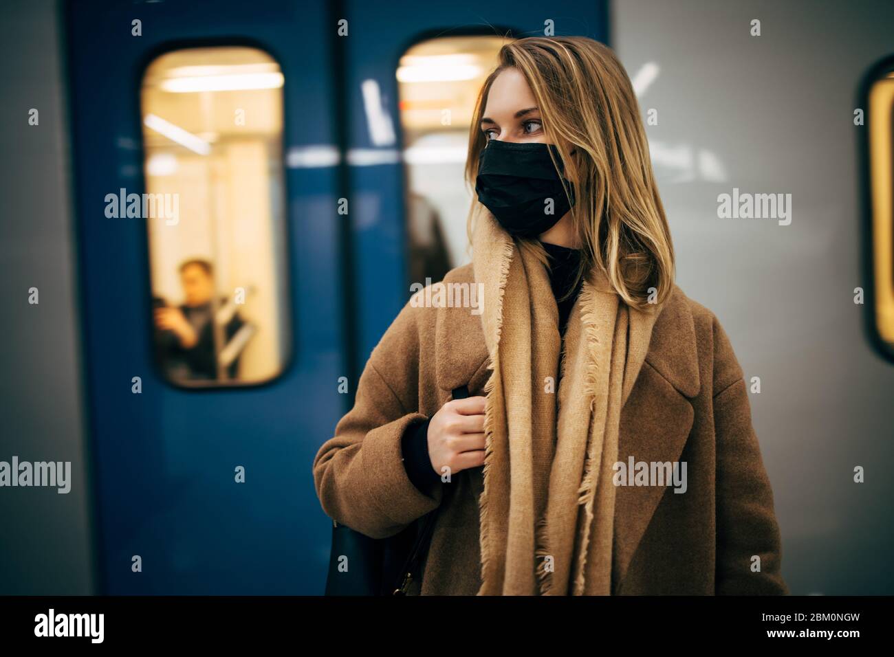 Blonde woman in black medical mask looking at side while standing in subway near carriage. Coronavirus epidemic. Stock Photo