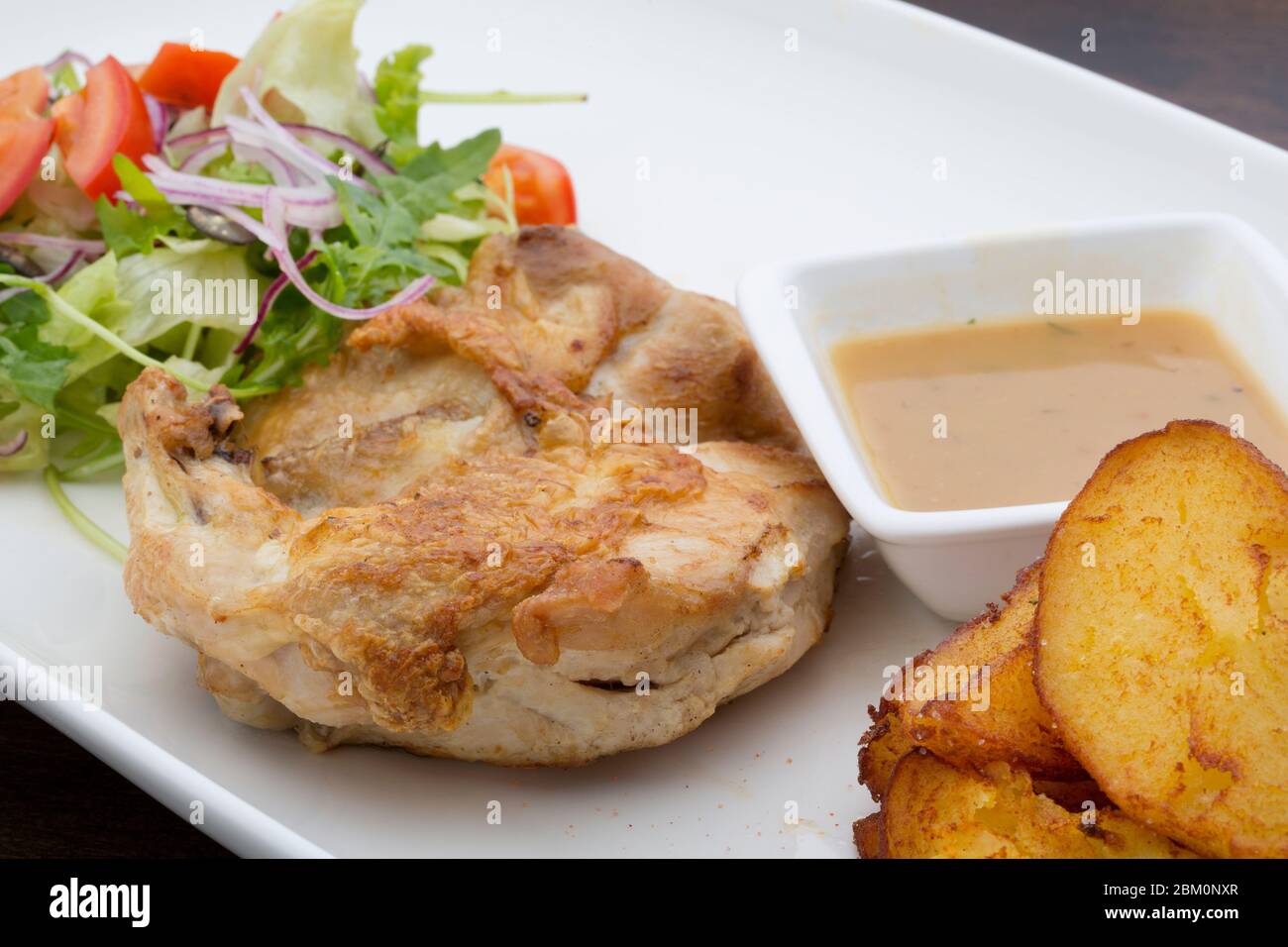 Roasted chicken with sauce, baked potatoes and a mixed salad in a white ceramic plate on a wooden table Stock Photo
