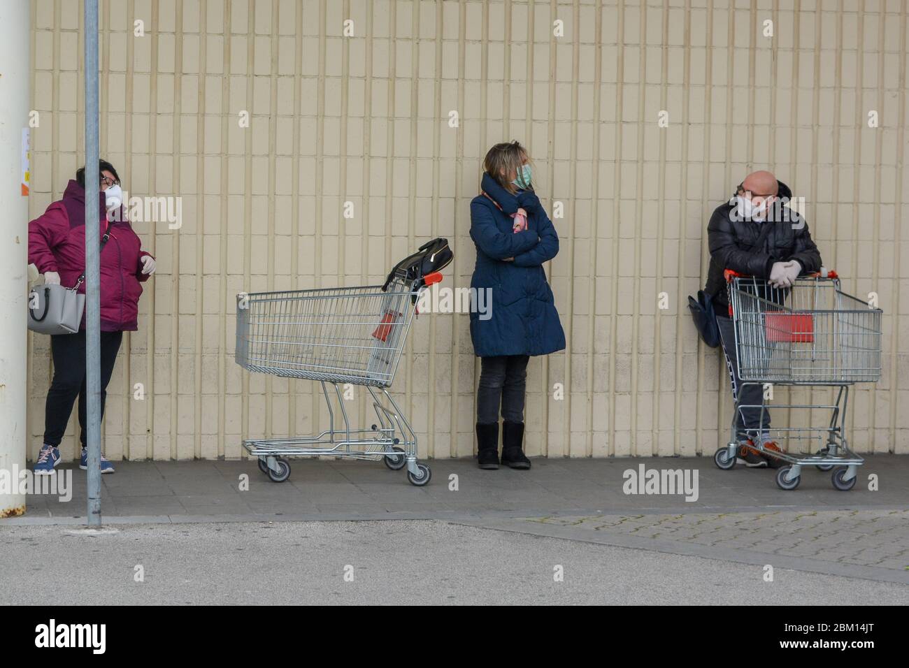 Toscana, Italy 03/27/20 - Customers wearing coronavirus protection masks in line with shopping carts, standing against a white wall. People chatting Stock Photo
