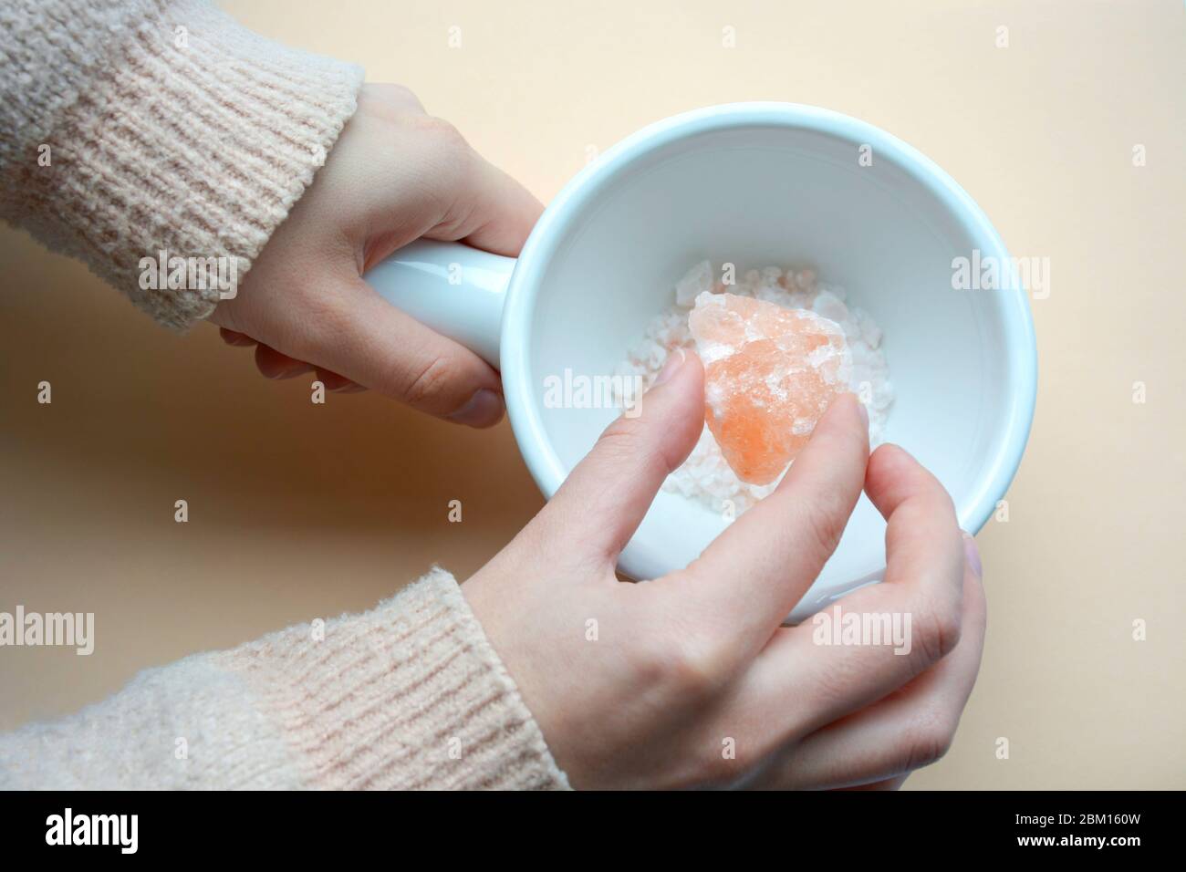 Female hand holding a Himalayan coral pink rock salt crystal above a ceramic white bowl on coral background Stock Photo