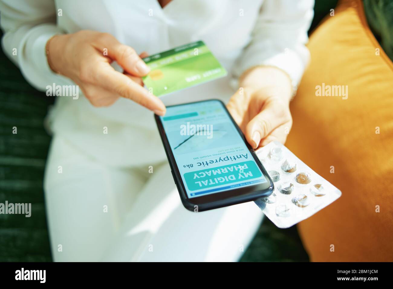 Upper view of female with credit card and empty blister pack of pills using online pharmacy store on a smartphone in the living room in sunny day. Stock Photo