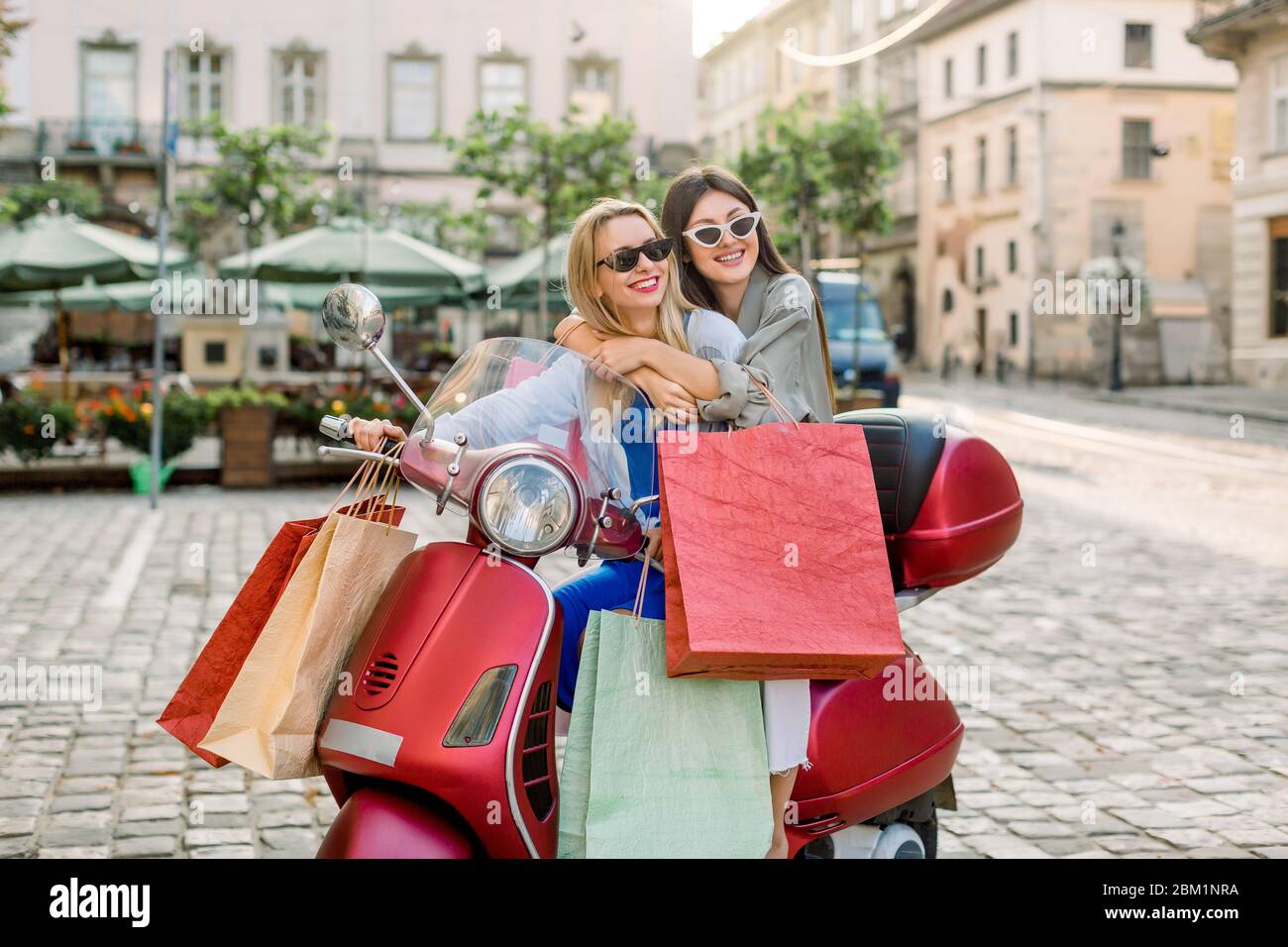 Two attractive young girls best friends wearing casual clothes and  sunglasses sitting on the red scooter with a lot of shop bags, on the  background of Stock Photo - Alamy