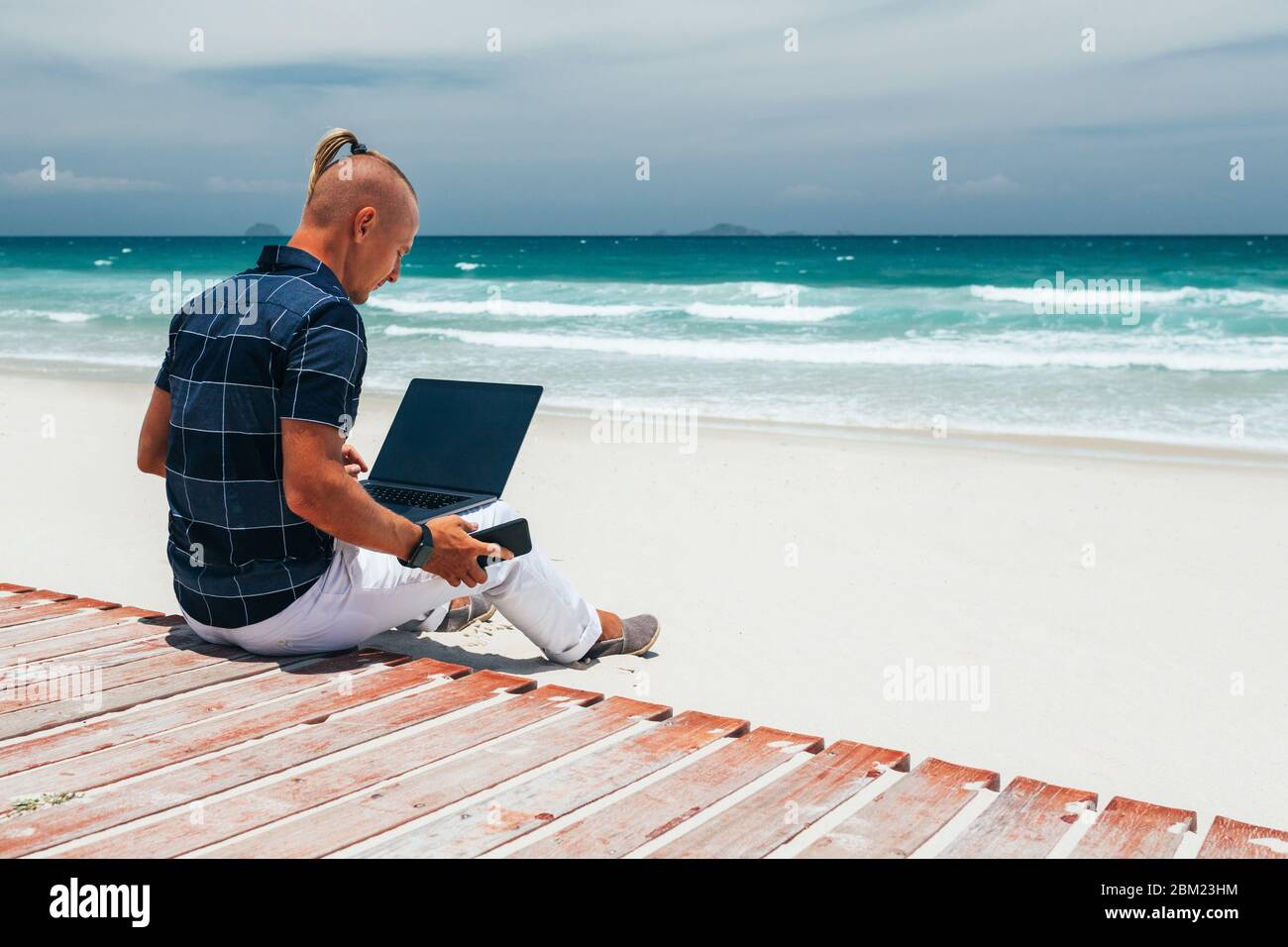 Young guy Holds smartphone in hand. working using a laptop, sitting on the seashore on a sandy beach. Businessman  on a journey. freelancer makes a co Stock Photo