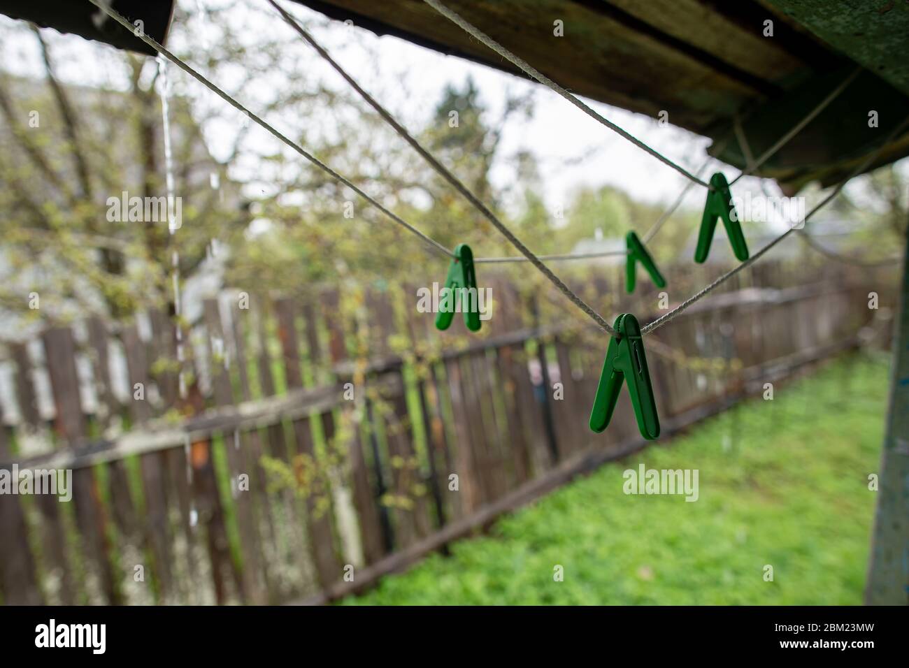 Plastic clothespins hang on a clothesline, under the porch roof, in the rain, against the background of an old fence and garden. Stock Photo