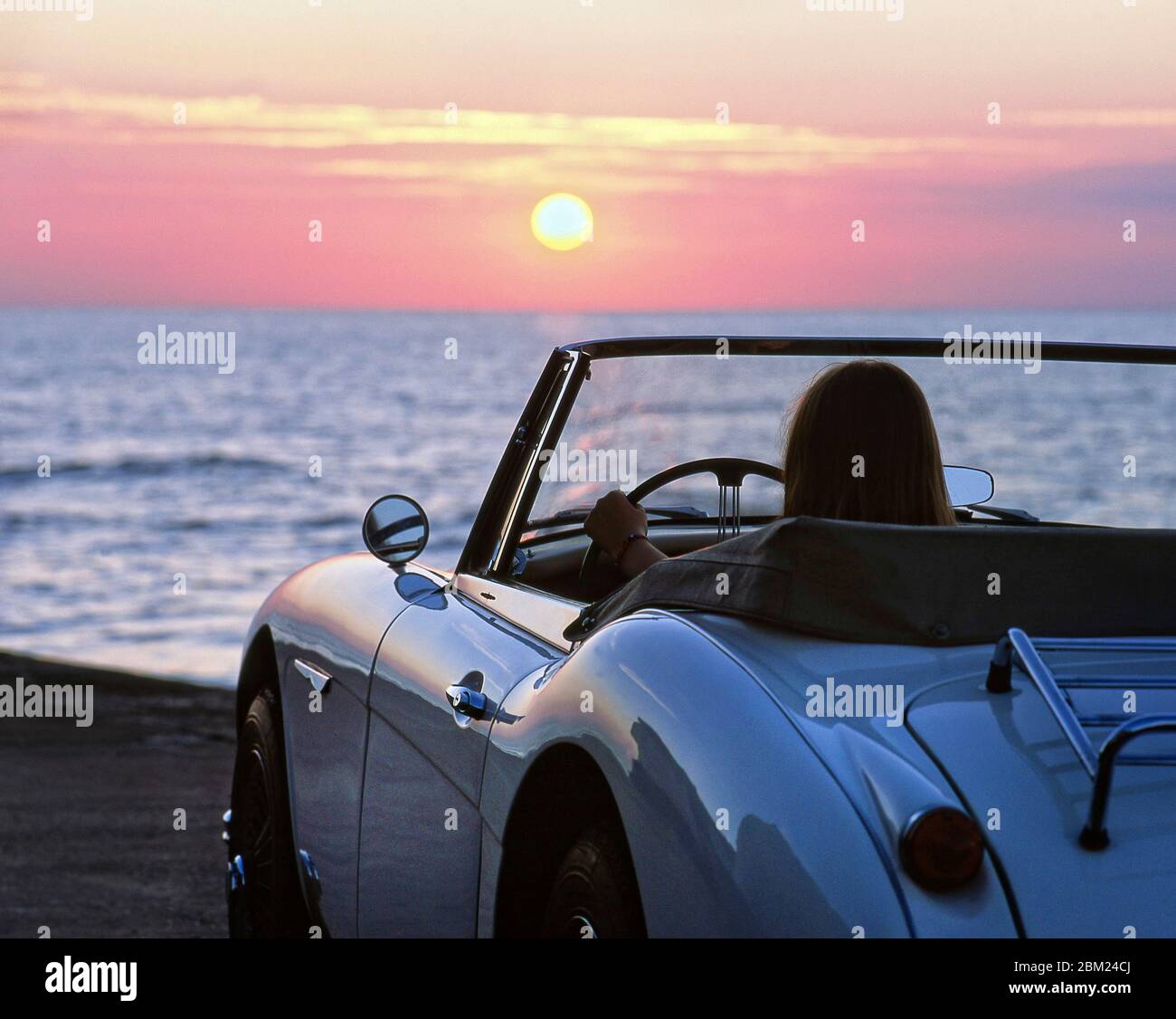 Young Woman driving a Left hand drive 1965 Austin Healey 3000 at sunset on coastal road in Kent UK Stock Photo
