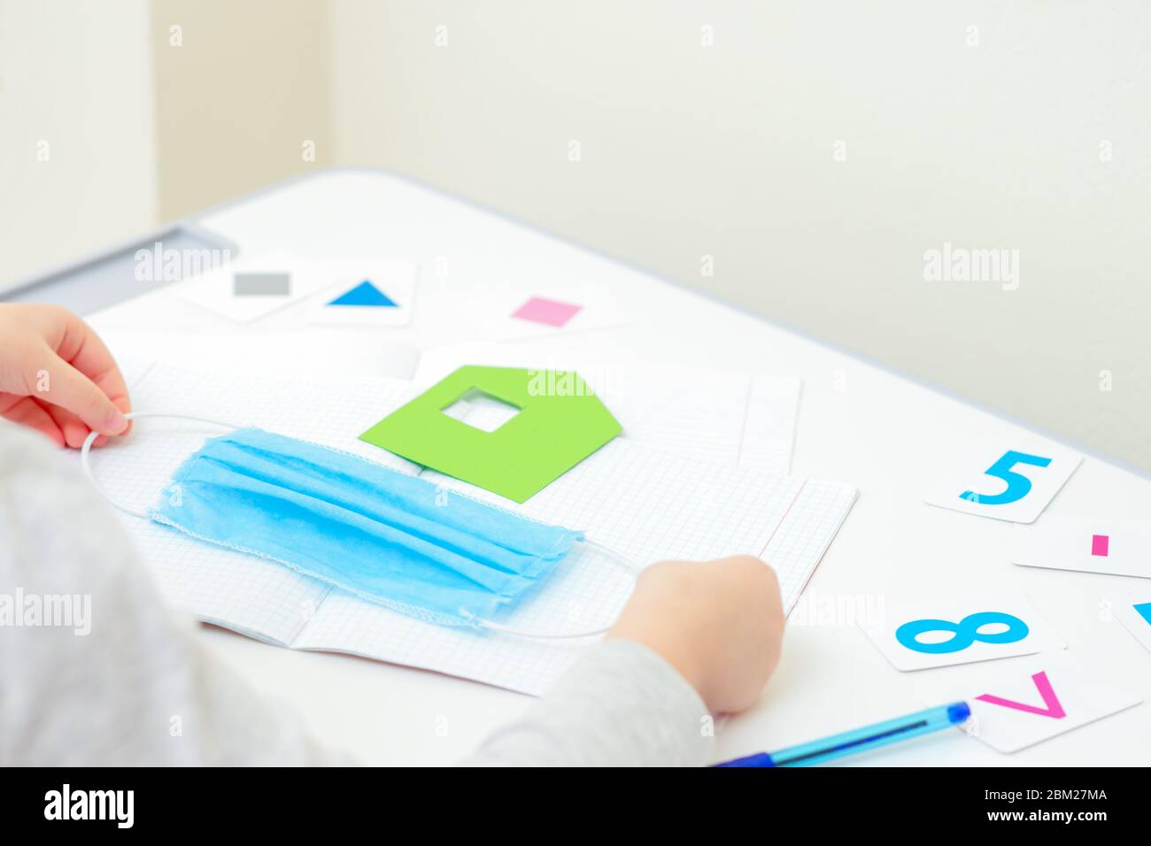 Little girl is holding medical mask near paper green house over copybook with numbers on white desk at home. Coronavirus outbreak concept. Stock Photo