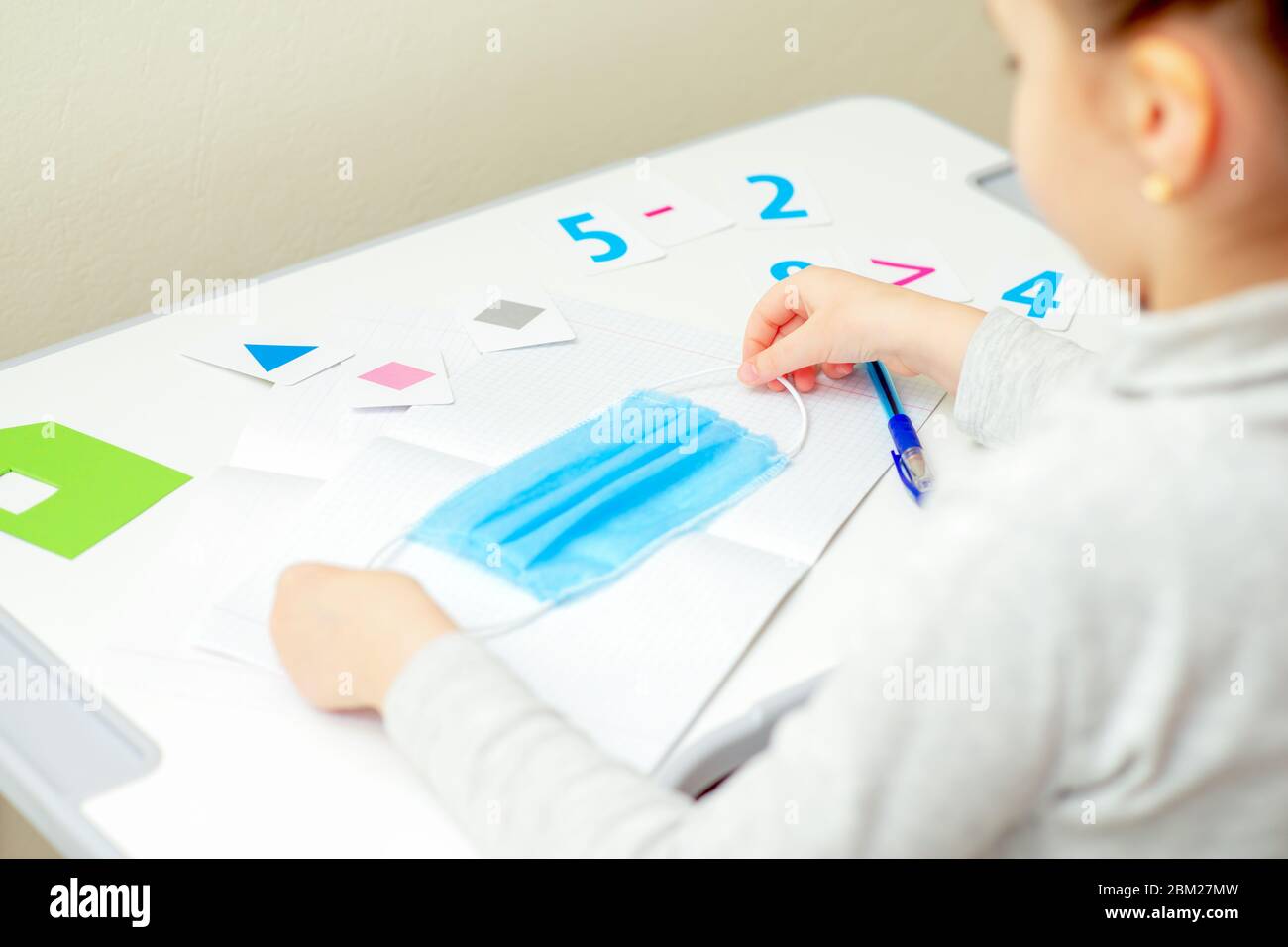 Little girl is holding medical mask for protection over copybook on white desk at home. Quarantine studying concept. Stock Photo