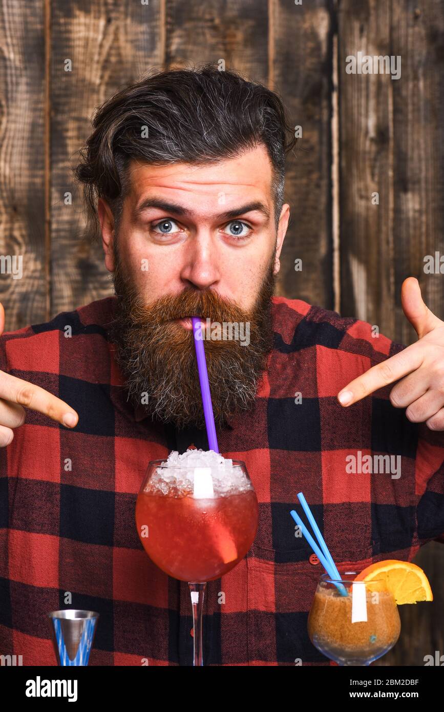 Hipster enjoy drink or cocktail. Barman with beard and surprised face drinks out of glass with drinking straw cocktail. Man in checkered shirt on wooden background. Alcohol and cocktails concept Stock Photo
