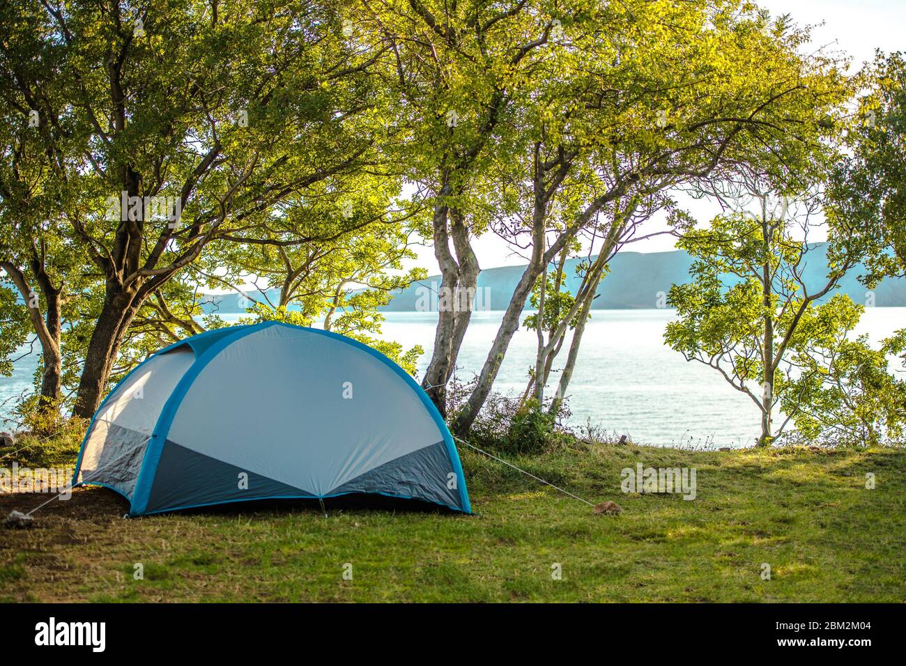 View Of Blue Small Tent Pitched On Shaded By Trees Area Near Lake And Mountains. Stock Photo