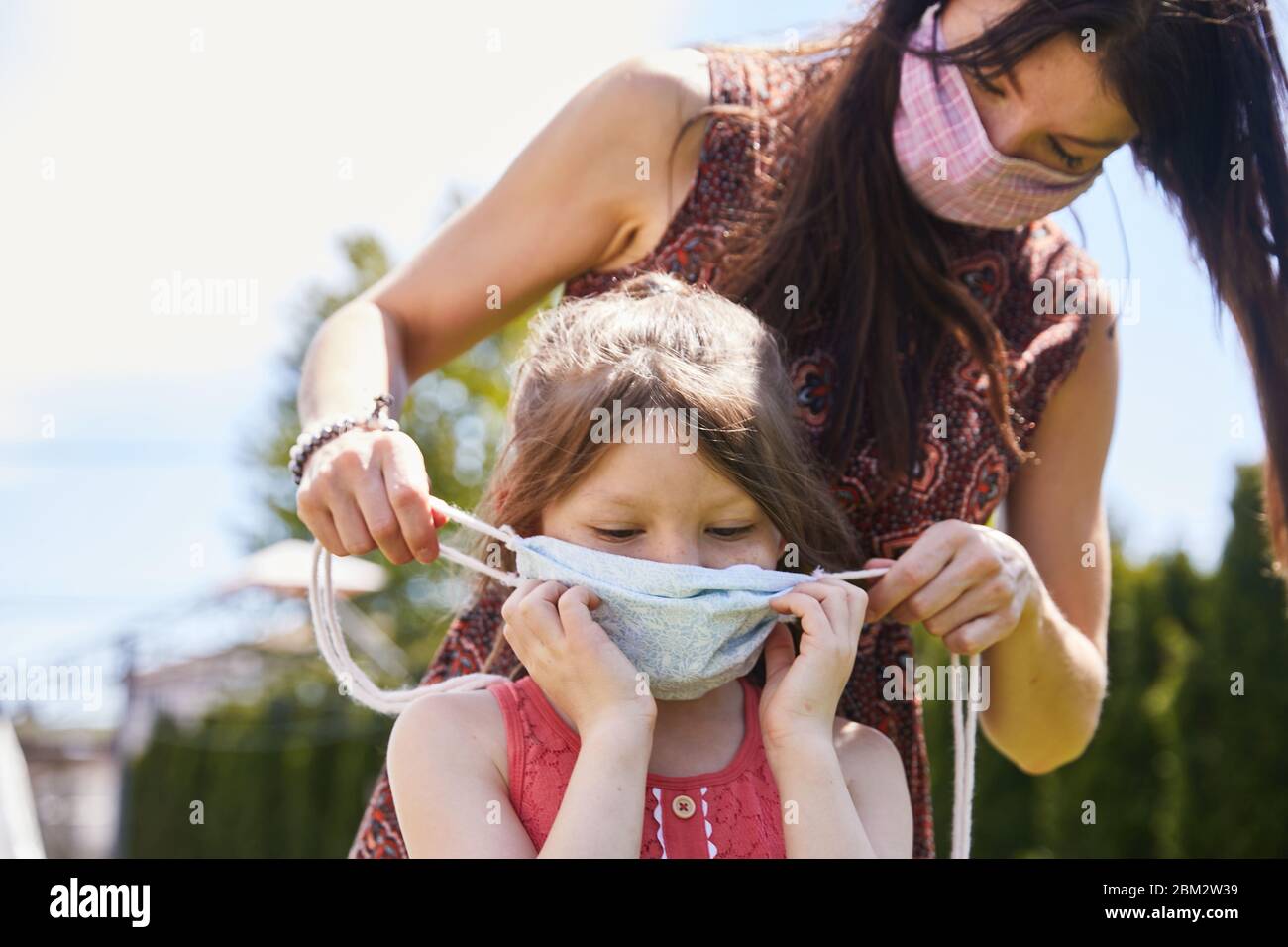 A mother lovingly ties a facemask on her daughter outdoors at a park to safeguard against infection from covid19 Stock Photo