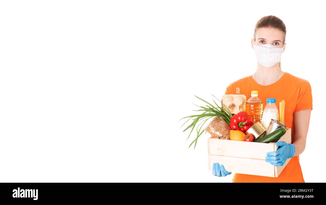 young girl in a mask and an orange t-shirt holds a wooden box with groceries for delivery isolated on white background Stock Photo