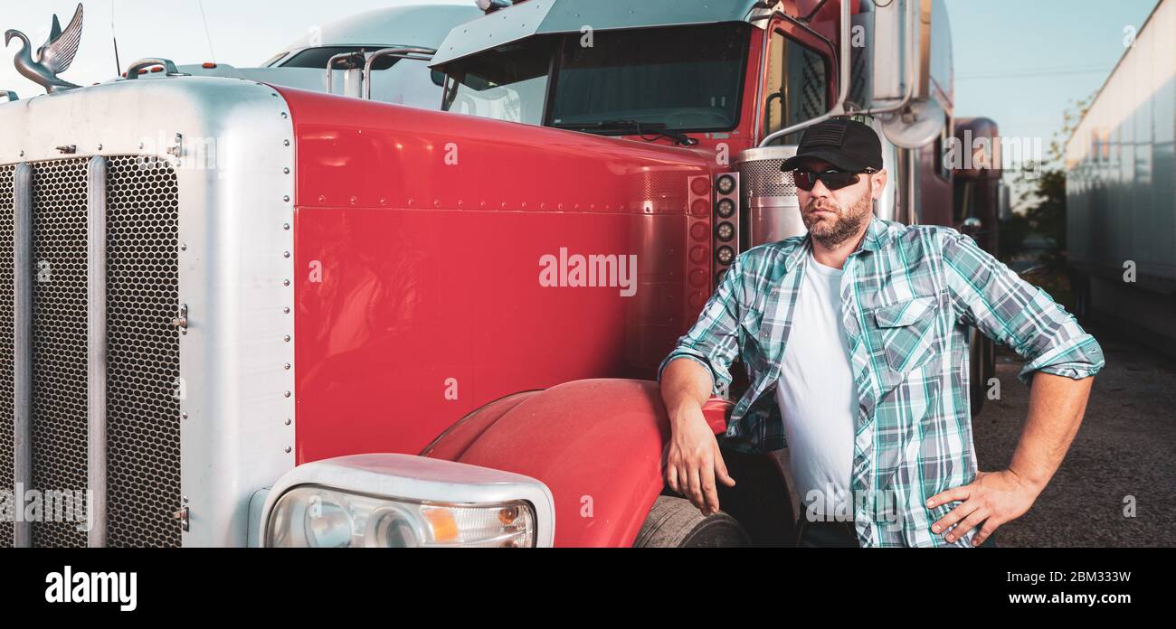 Semi truck professional driver on the job in casual clothing. Confident looking trucker stands next to red big rig wearing sunglasses and baseball cap Stock Photo