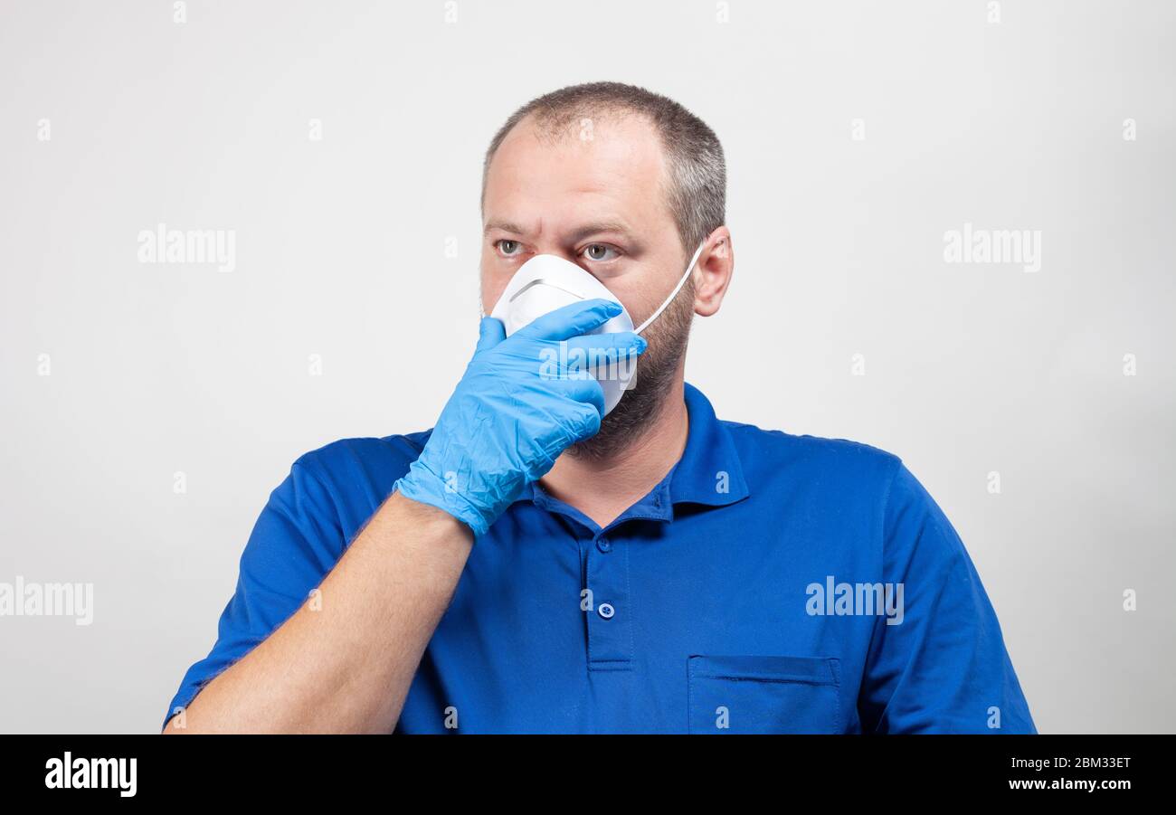 Portrait of man in his mid 30's equipped with protective mask and surgical mask on white. Healthcare worker holds hand over face mask. Stock Photo