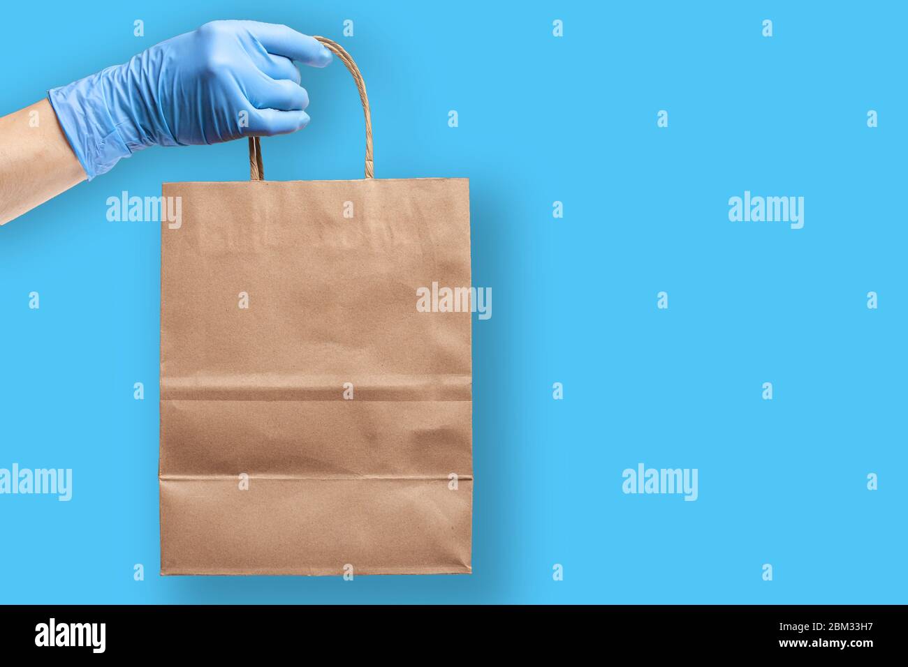 Hand wearing rubber protective glove   holds paper grocery bag. Grocery shopping and delivery concept during novel virus pandemic. Stock Photo
