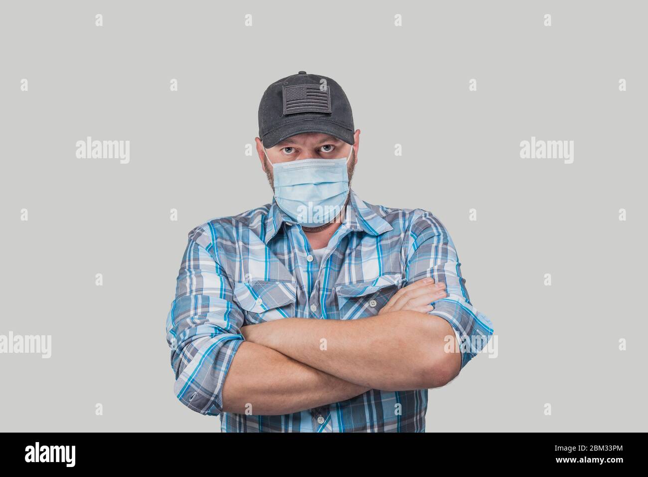 Caucasian man dressed casual with plain blue shirt and black baseball cap wears face medical hat and looks angrily at the camera. Stock Photo