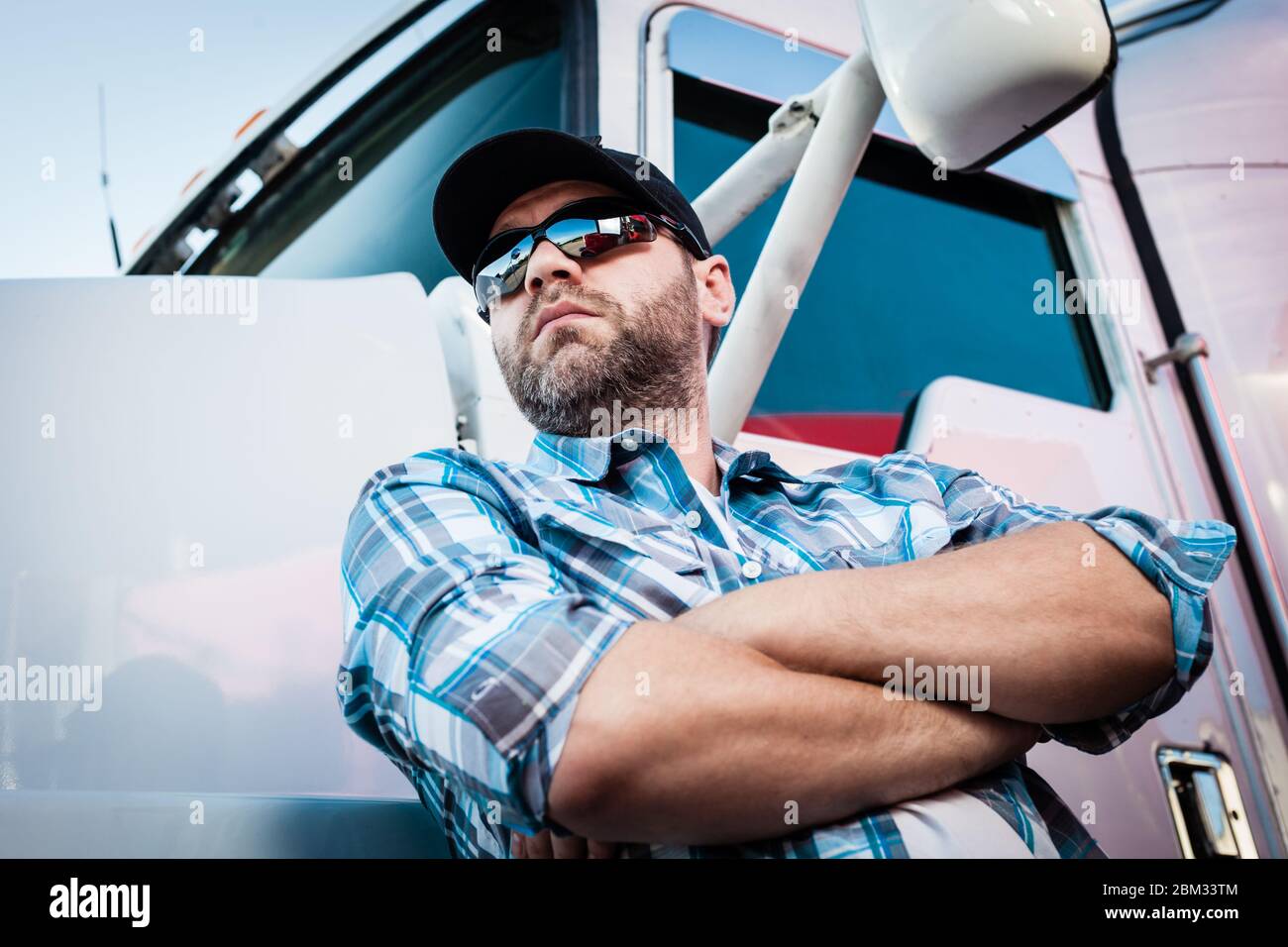 Confident Caucasian male american trucker next to his big rig. Concept of trucking owner operators with man in plaid shirt and baseball cap. Stock Photo