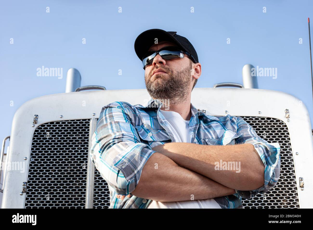 Confident semi truck driver wearing plaid shirt and black baseball cap stands with arms crossed in front of big rig. Stock Photo