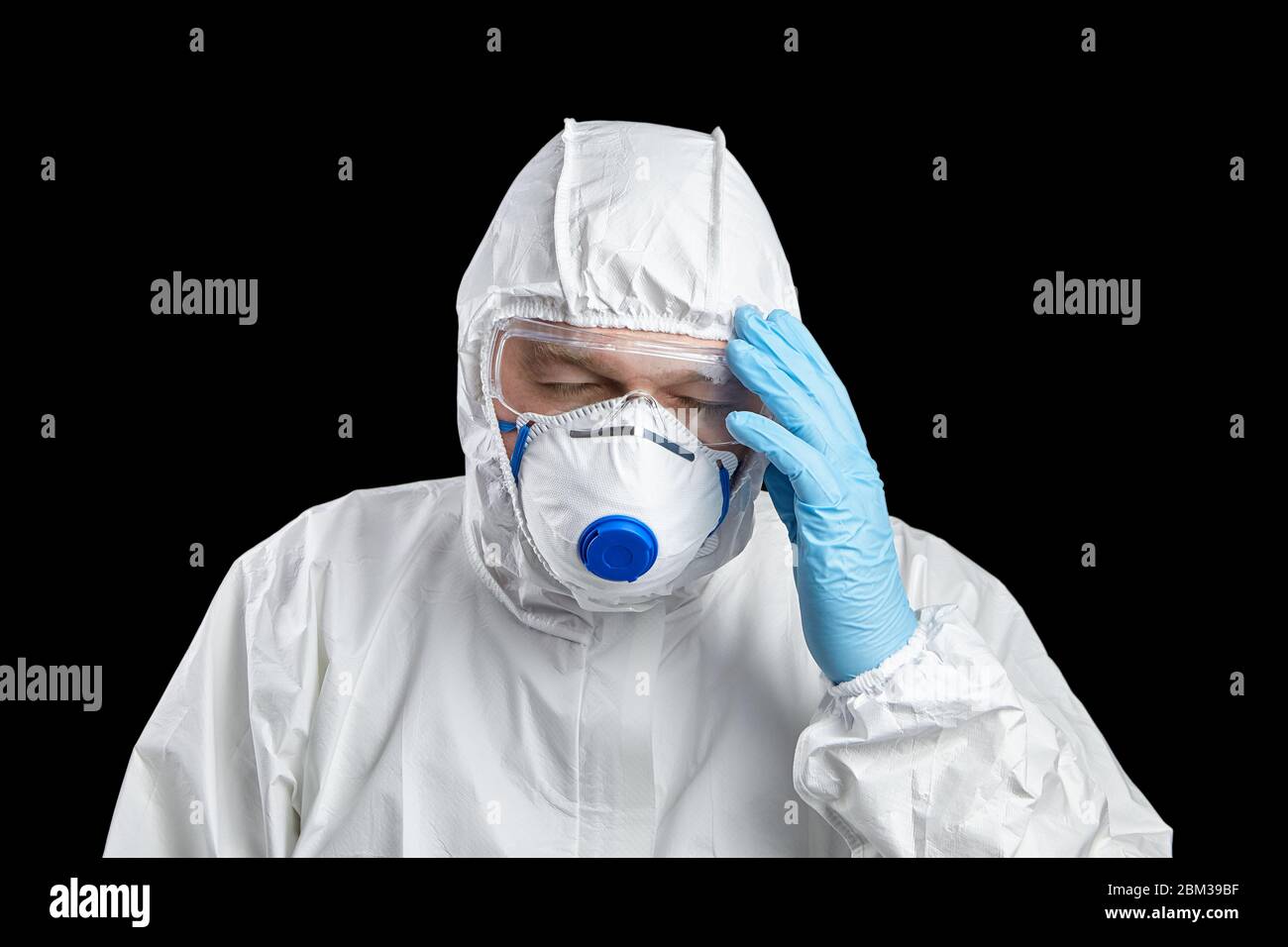 Tired doctor in white protective suit, facepiece respirator and goggles. Man holds his head, hand in disposable blue gloves, isolated on black backgro Stock Photo