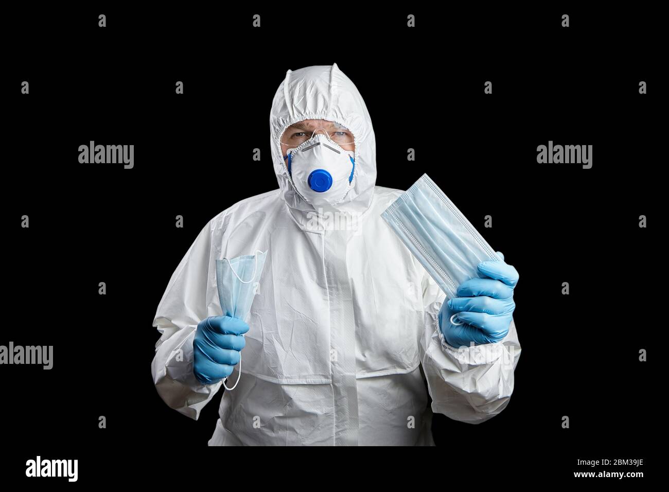 Doctor in white protective suit and facepiece respirator with medical face masks in hands, disposable blue gloves and googles, isolated on black backg Stock Photo