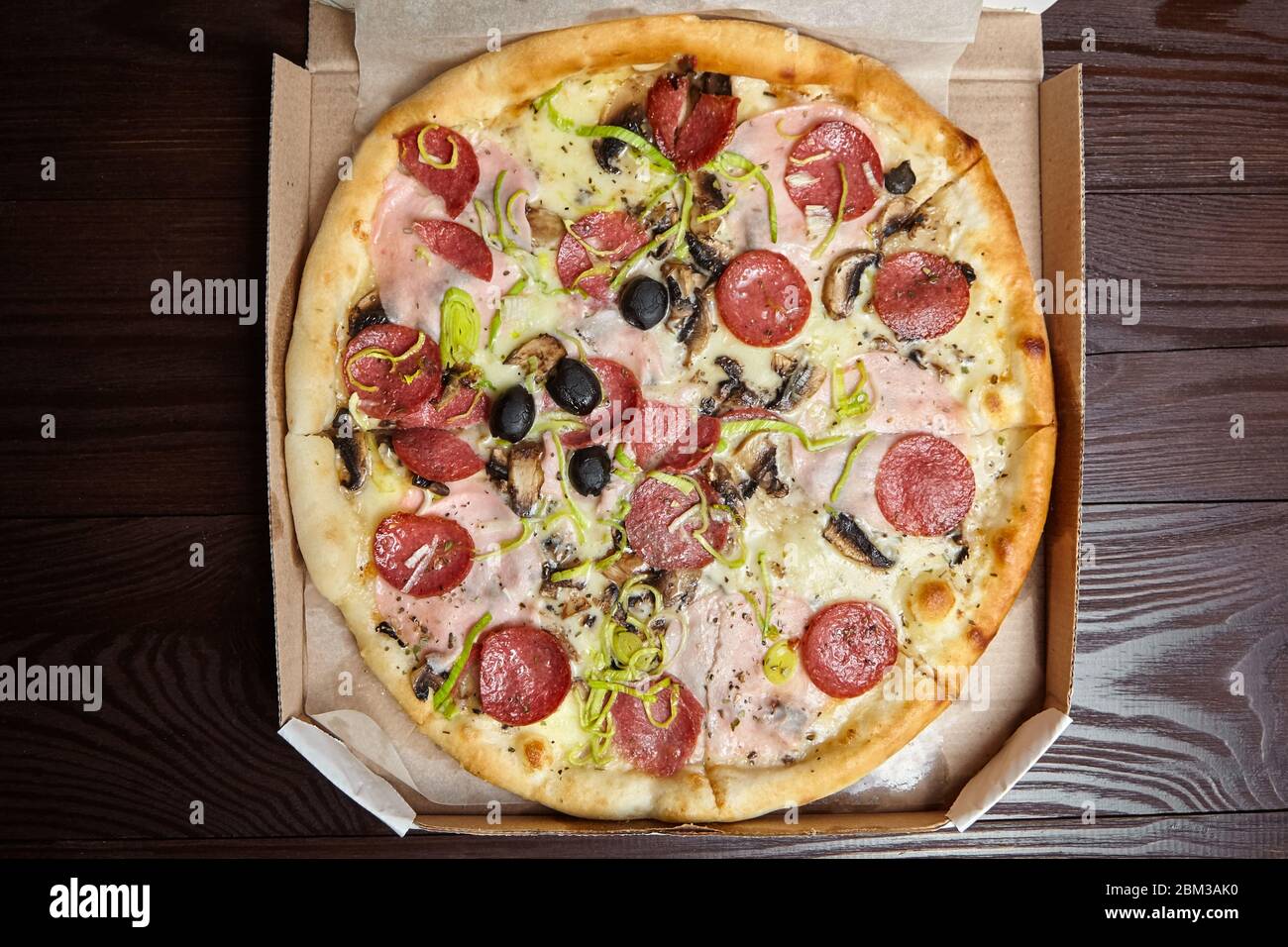 Pizza in a open cardboard box on brown wooden background, top view. Cooked pizza with white sauce, cheese, ham, salami, champignons, leek, black olive Stock Photo