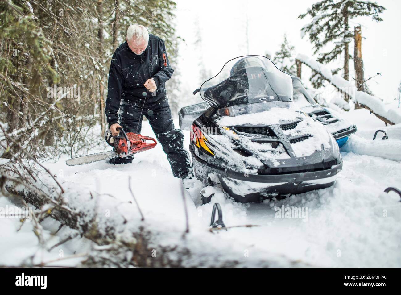 Retired man using chainsaw to clear trails while snowmobiling. Stock Photo