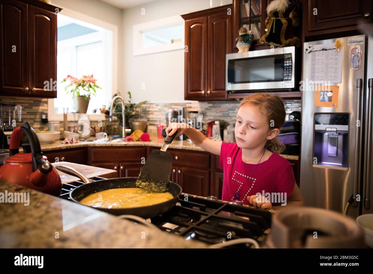Candid Image of Unsmiling Young Girl Cooking Eggs In Messy Kitchen Stock Photo