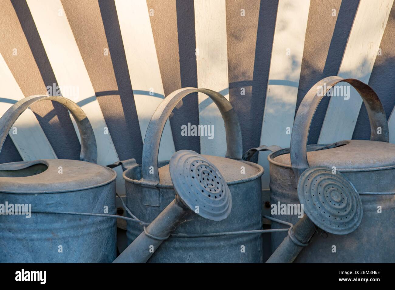 Old style galvanized iron watering cans lined up along a wall in the afternoon sun to form a pattern of sorts Stock Photo