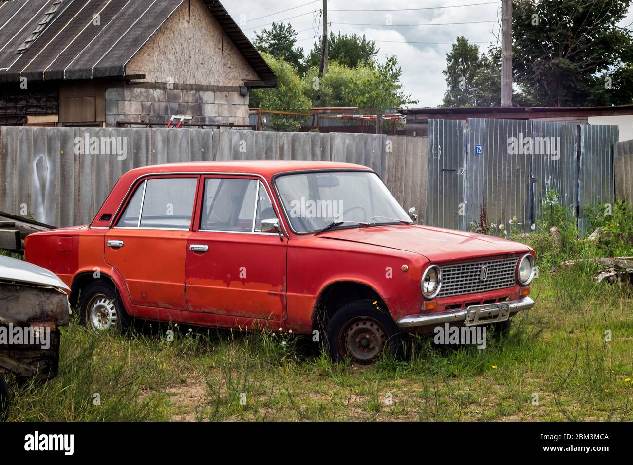 An old red Lada car with holey numbers is standing in front of a village house. Stock Photo