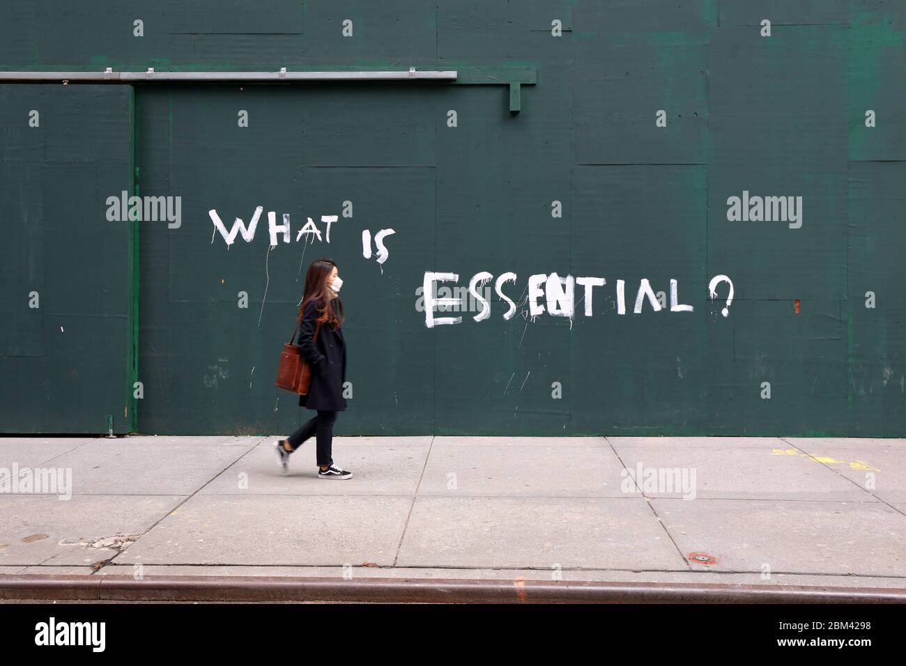 A person with a mask walks past a construction site with the words, "What is Essential?" painted on its walls, in New York, 27th April 2020 Stock Photo