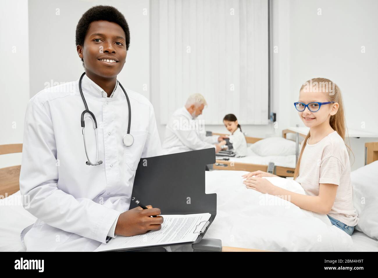 Young smiling african male doctor with stethoscope on neck and positive little patient sitting on coach in hospital, therapist holding folder and pen. Doctor measuring blood pressure on background. Stock Photo