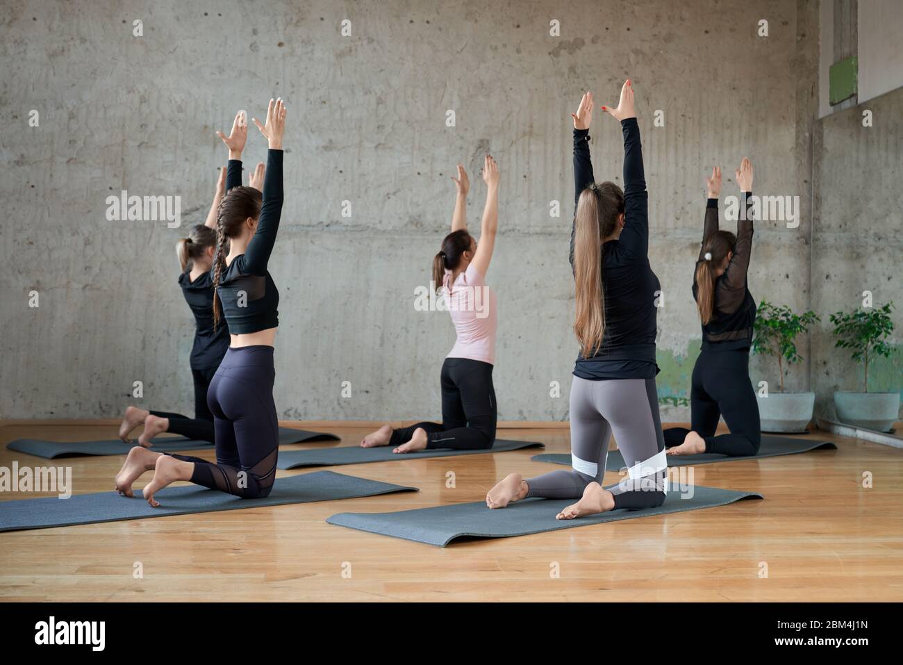Back view of young fitnesswomen in sportswear practicing yoga pose, standing on knees with hands up, practicing in studio, loft interior. Concept of healthy lifestyle, yoga. Stock Photo