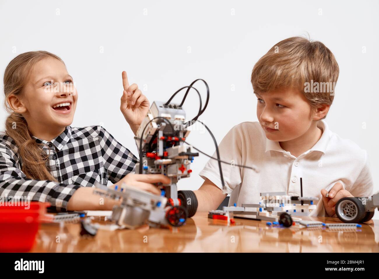 Front view of boy and girl having fun, creating vehicle. Science engineering. Friends working on project together, girl having idea and pointing with finger up. Building kit for kids on table. Stock Photo