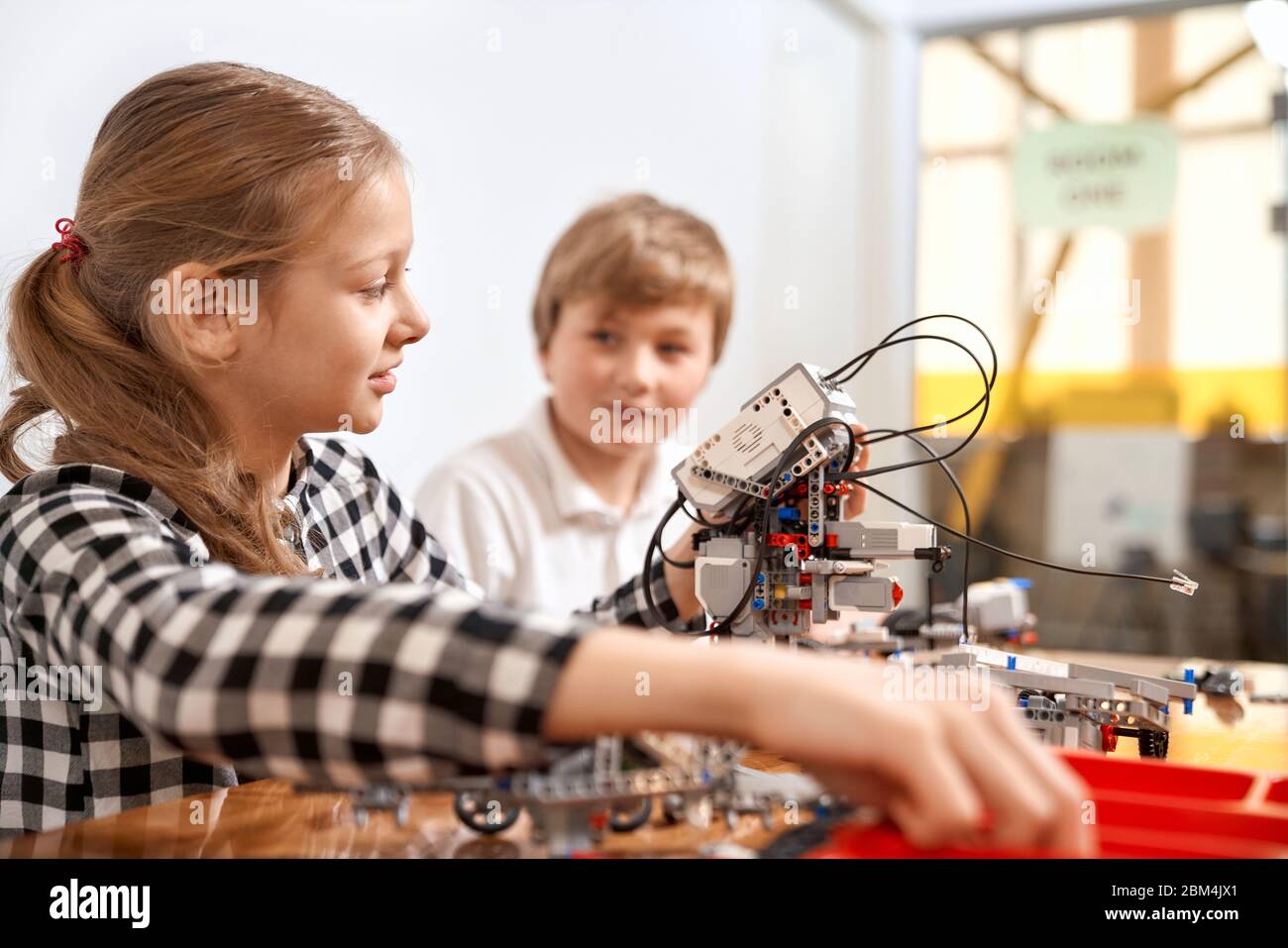 Side view of boy helping girl in creating robot using building kit for kids on table. Science engineering. Nice interested friends smiling, chatting and working on project together. Stock Photo
