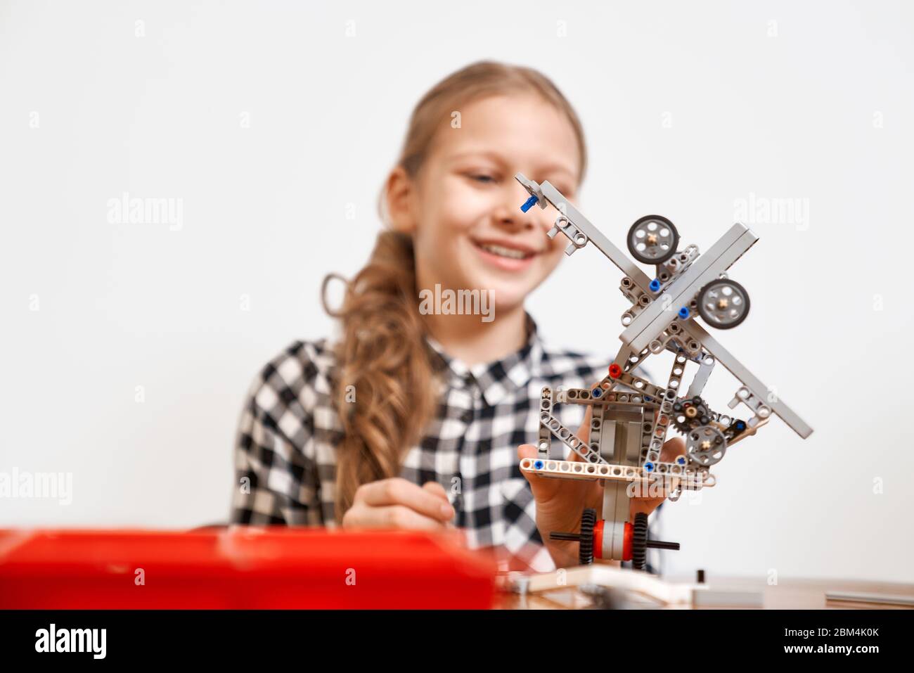Selective focus of interesting robot in hands of young smiling girl sitting at table. Close up of vehicle made using building kit fo children. Concept of science engineering. Stock Photo