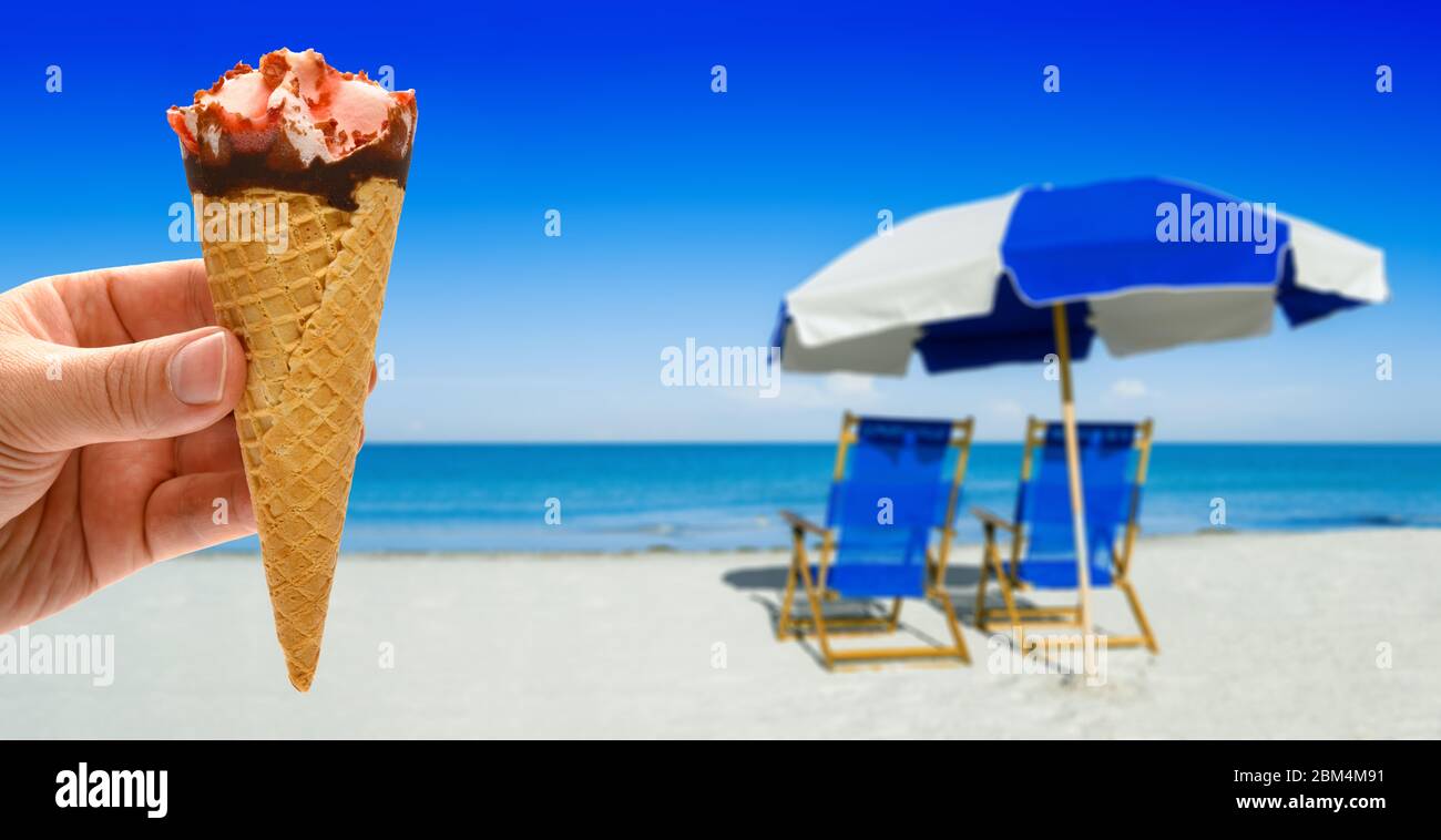 hand holding a fresh strawberry flavor ice cream cone in front of blurred sun loungers and a beach umbrella on silver sand Stock Photo