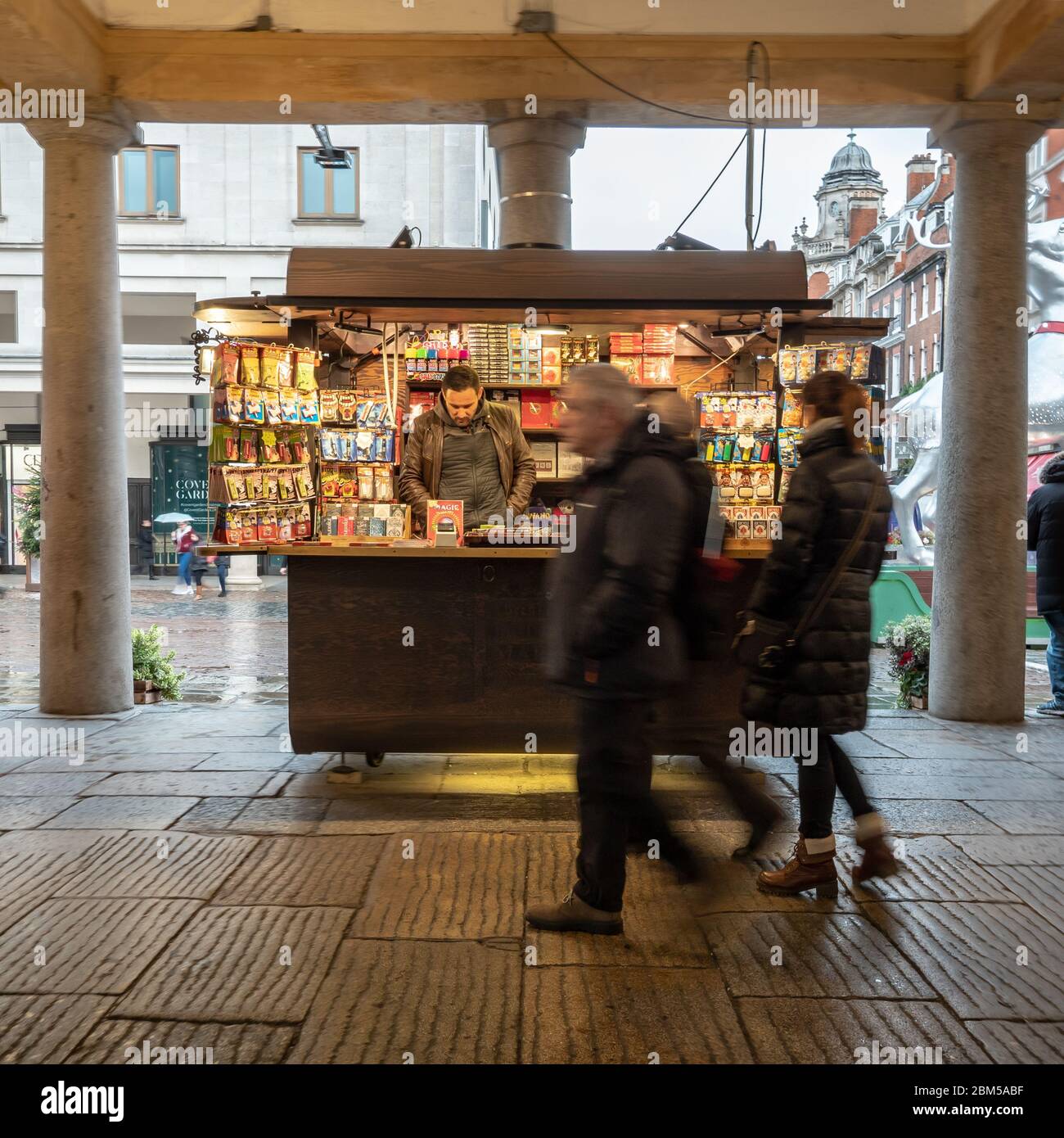 Covent Garden street trader. Candid shoppers walking by a brightly lit market stall holder in the heart of London's West End tourist district. Stock Photo