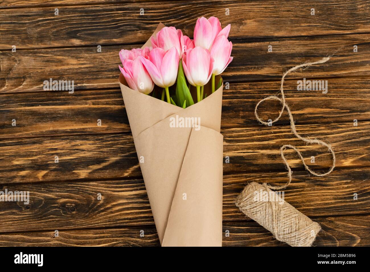 top view of pink tulips wrapped in paper near jute twine rope on wooden ...