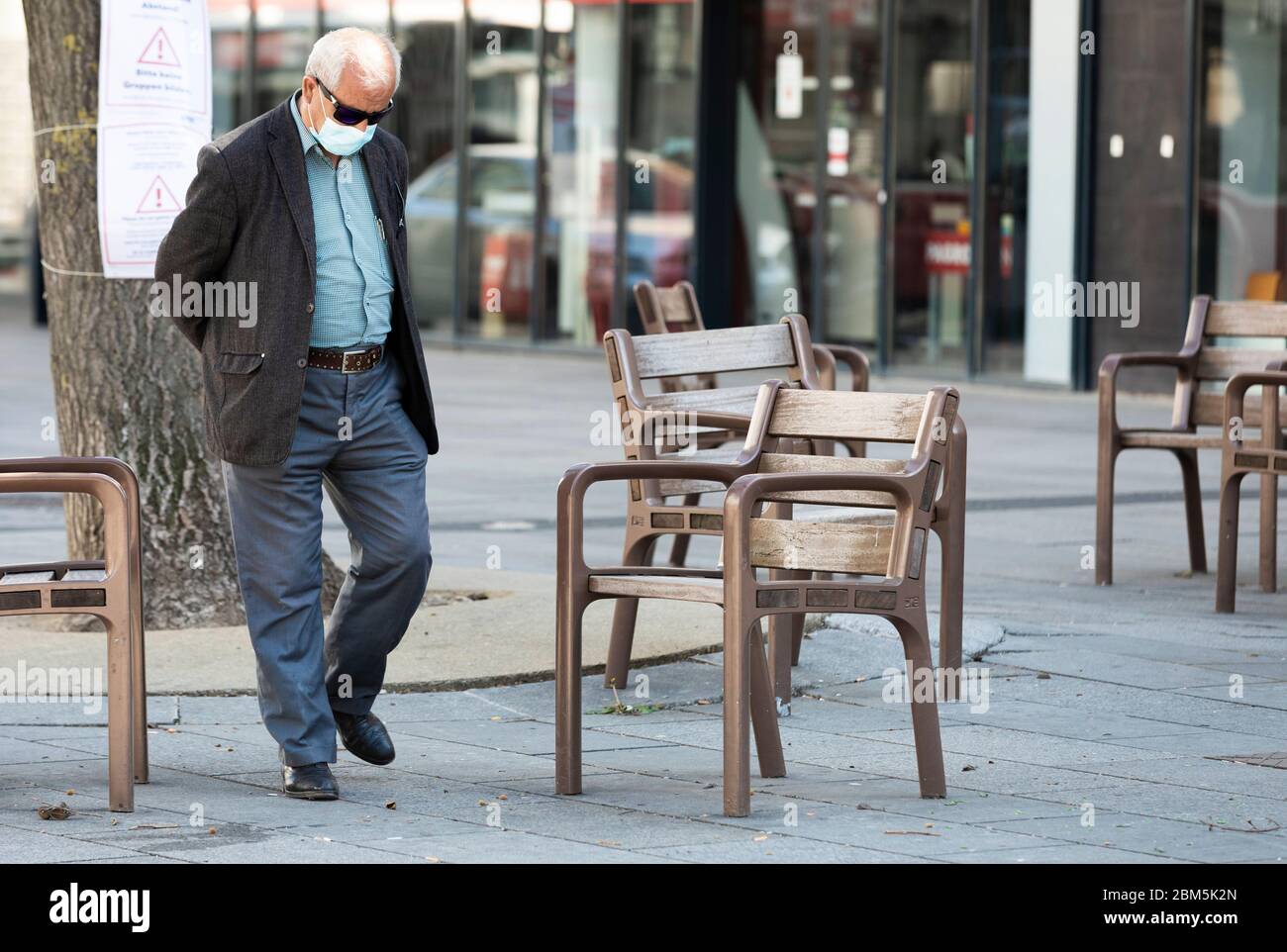 old man with a coronavirus protection mask walking on a square with empty chairs Stock Photo